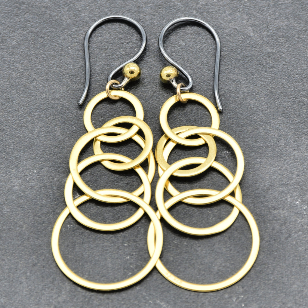 A pair of gold-toned dangle earrings with stacked interlocking circular rings and blackened silver ear wires, displayed on a dark gray textured background - made with love by Ruby Acorn.