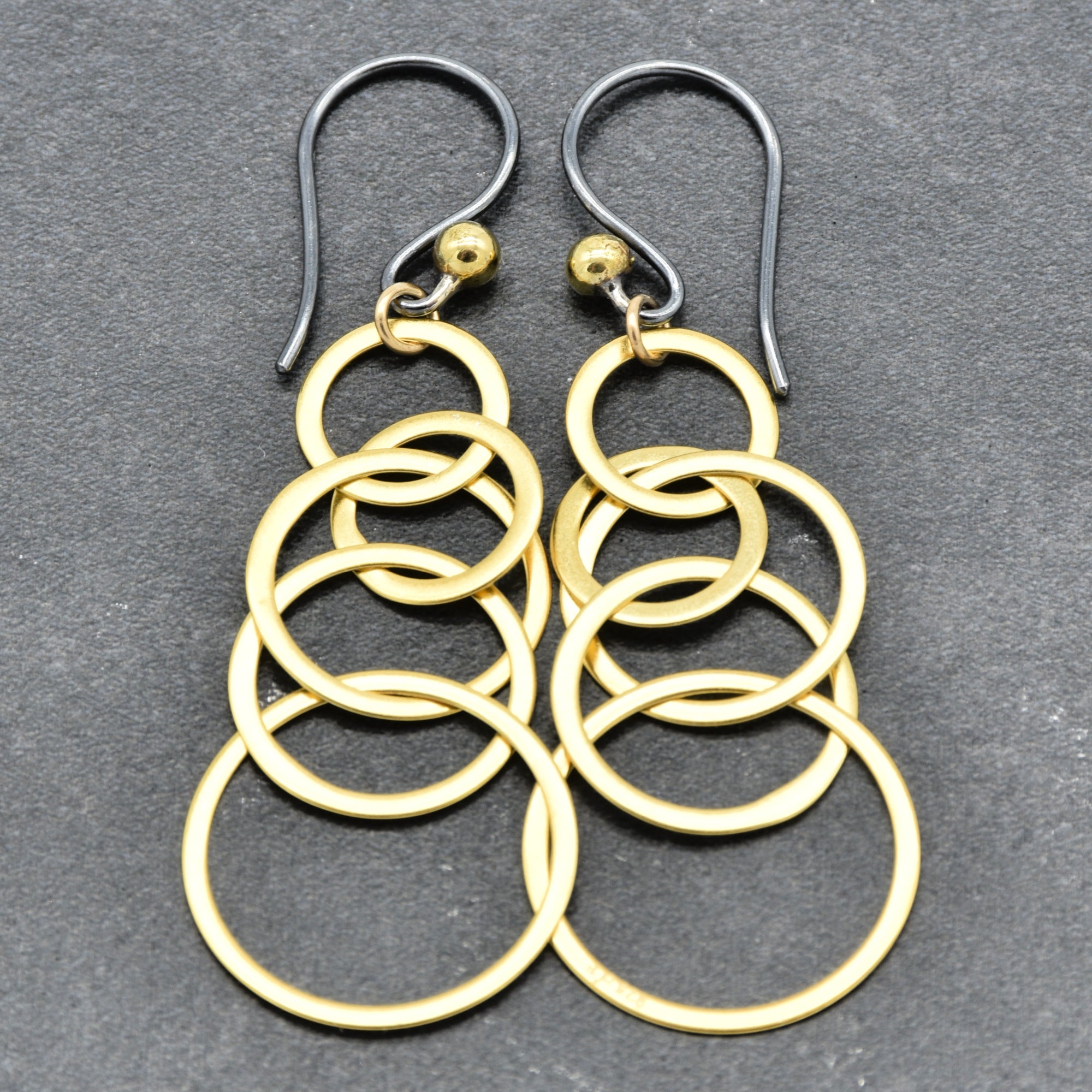 A pair of gold-toned dangle earrings with stacked interlocking circular rings and blackened silver ear wires, displayed on a dark gray textured background - made with love by Ruby Acorn.