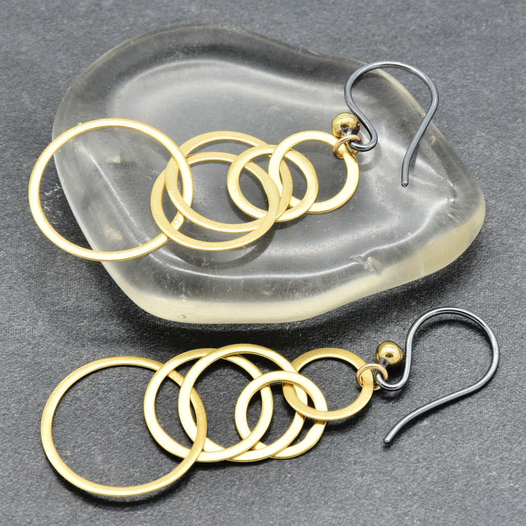 A pair of gold and silver dangle earrings with five interlocking circular rings, displayed on a translucent glass dish and a dark gray textured surface - made with love by Ruby Acorn.