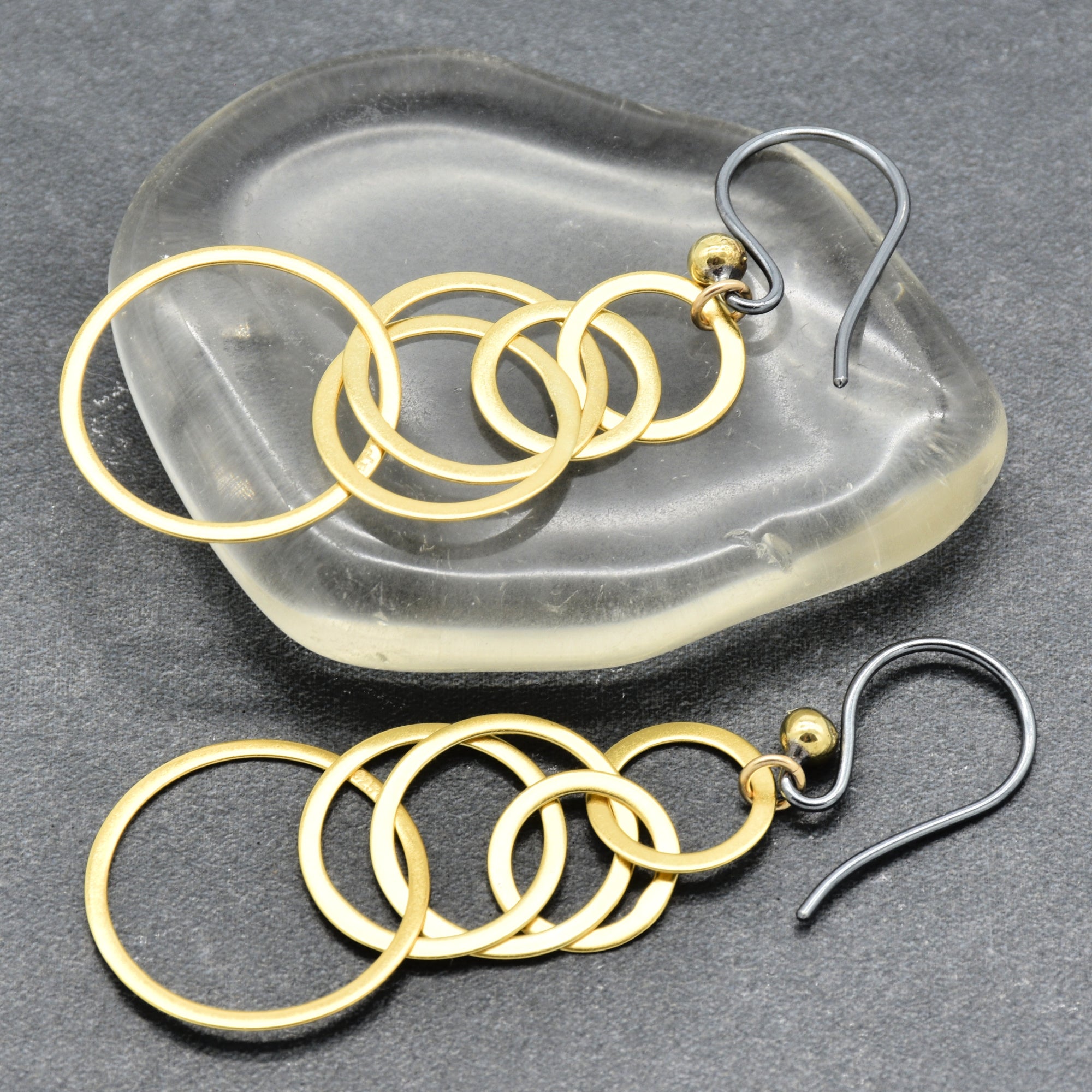 A pair of gold and silver dangle earrings with five interlocking circular rings, displayed on a translucent glass dish and a dark gray textured surface - made with love by Ruby Acorn.