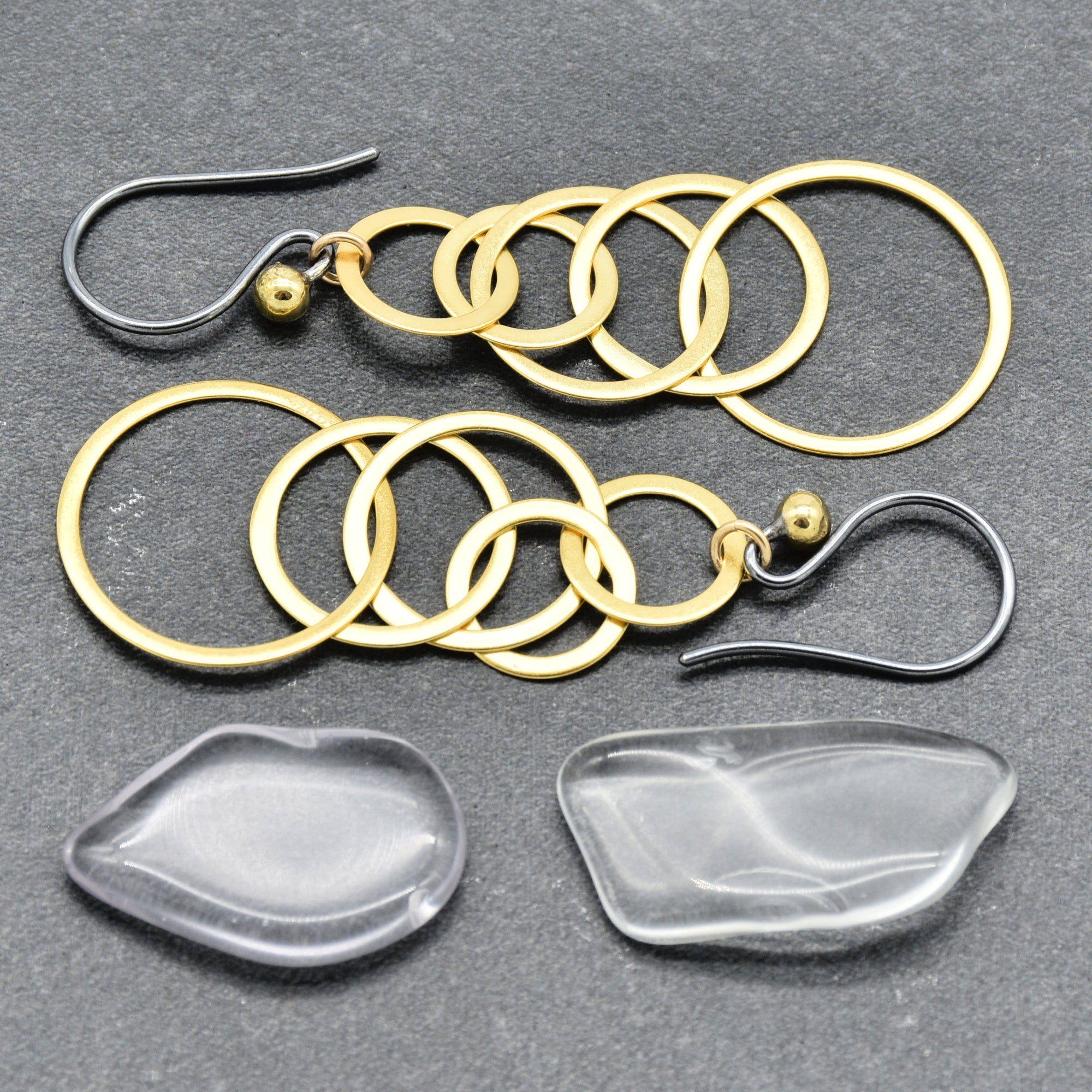 Handmade dangle earrings displayed on a dark gray textured background, alongside two flat, polished clear stone drops. The earrings feature multiple interlocking gold-tone rings, small gold beads, and black silver hooks - made with love by Ruby Acorn.