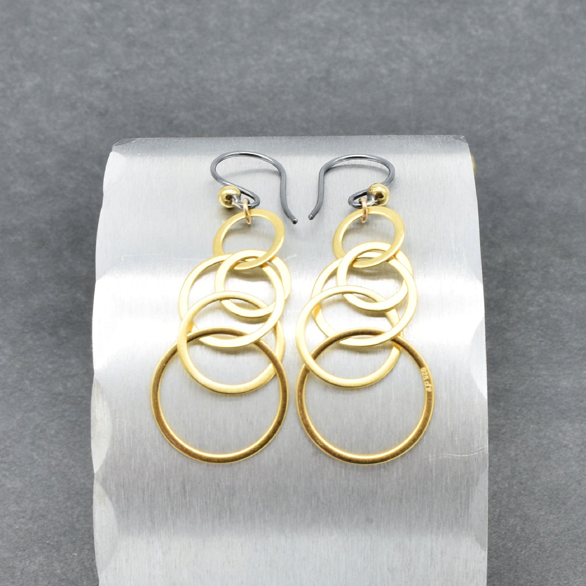 Gold interlocking hoop earrings with black silver ear wires on a silver circular display - made with love by Ruby Acorn.