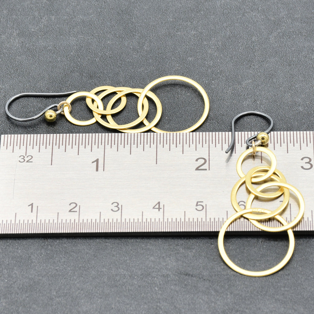 A pair of gold-toned dangle earrings made of interlocking circular rings, shown next to a metal ruler on a dark gray textured background to indicate scale - made with love by Ruby Acorn.