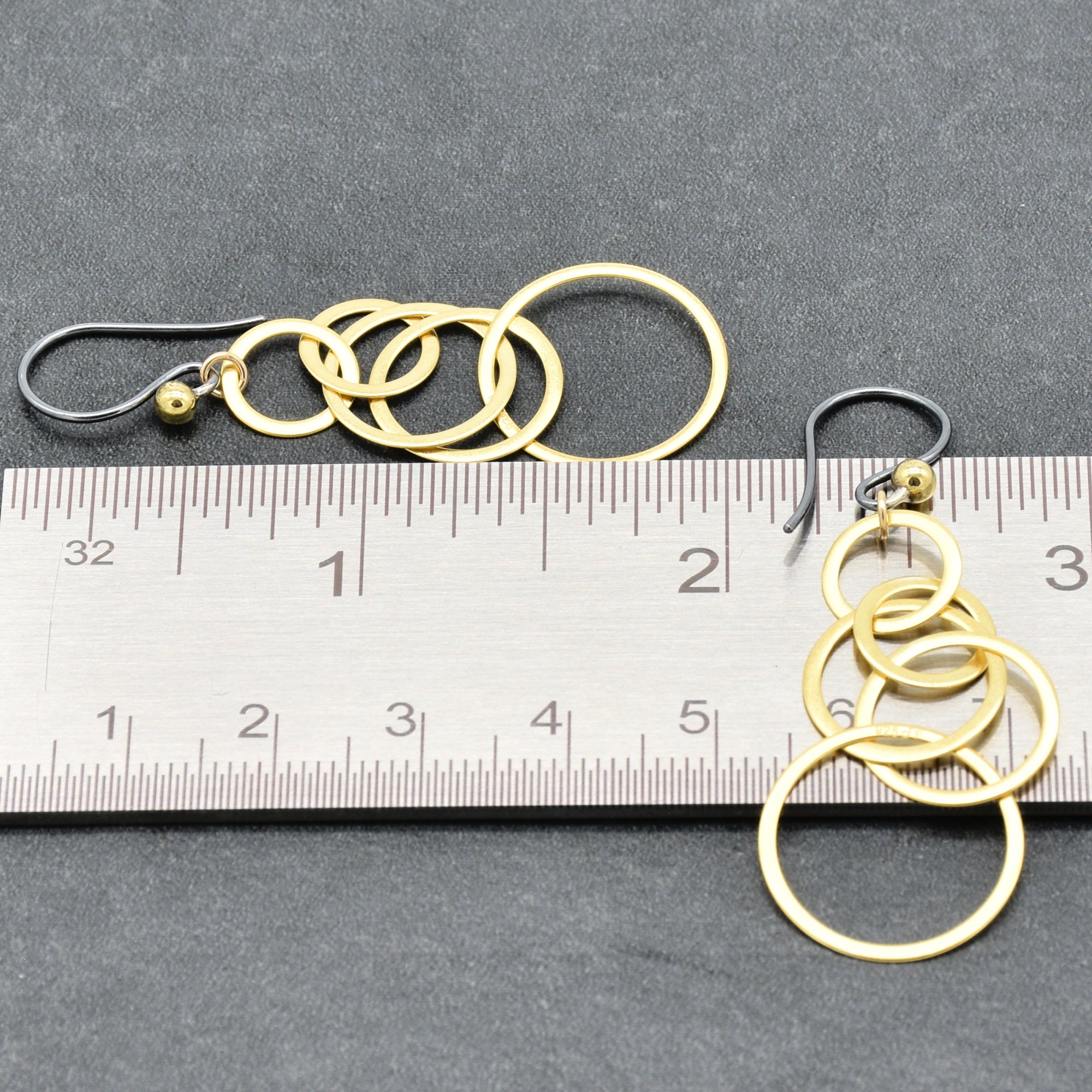 A pair of gold-toned dangle earrings made of interlocking circular rings, shown next to a metal ruler on a dark gray textured background to indicate scale - made with love by Ruby Acorn.