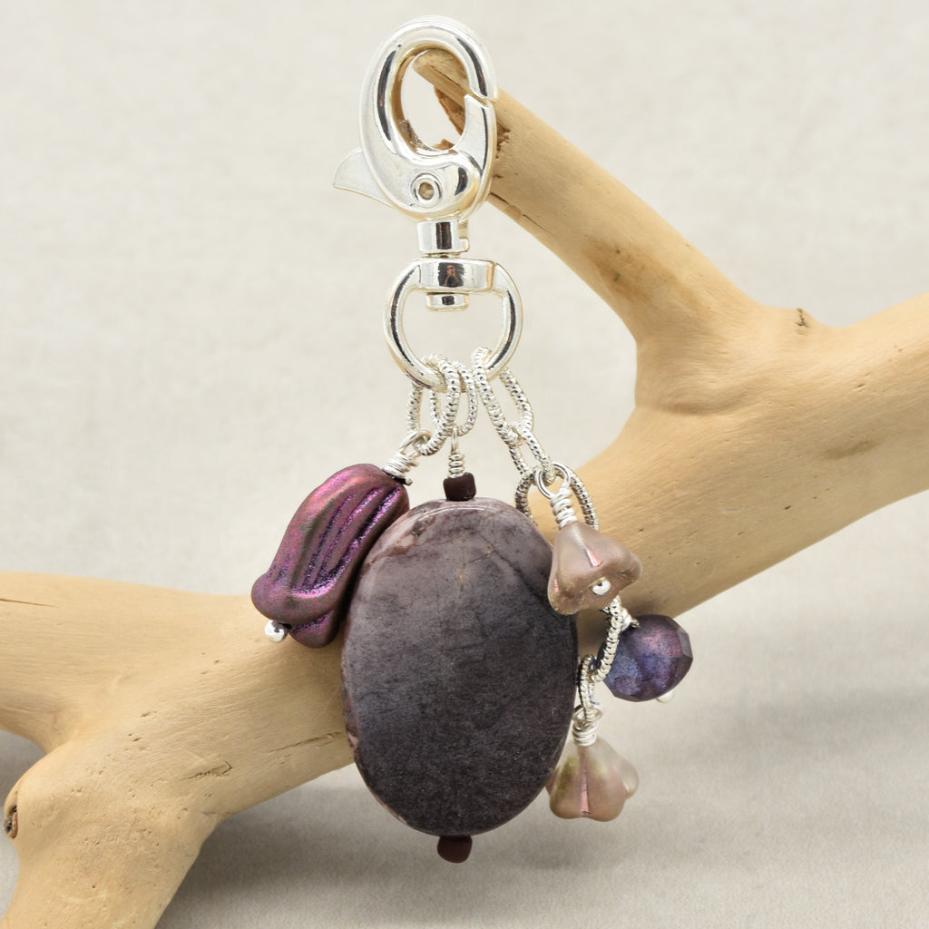 A handmade keychain featuring an oval porcelain jasper focal, a metallic purple Czech glass tulip charm, a small taupe flower charms, and a purple bead, all attached to a silver lobster clasp and displayed on a light wood branch against a neutral beige background - made with love in Colorado by Ruby Acorn.