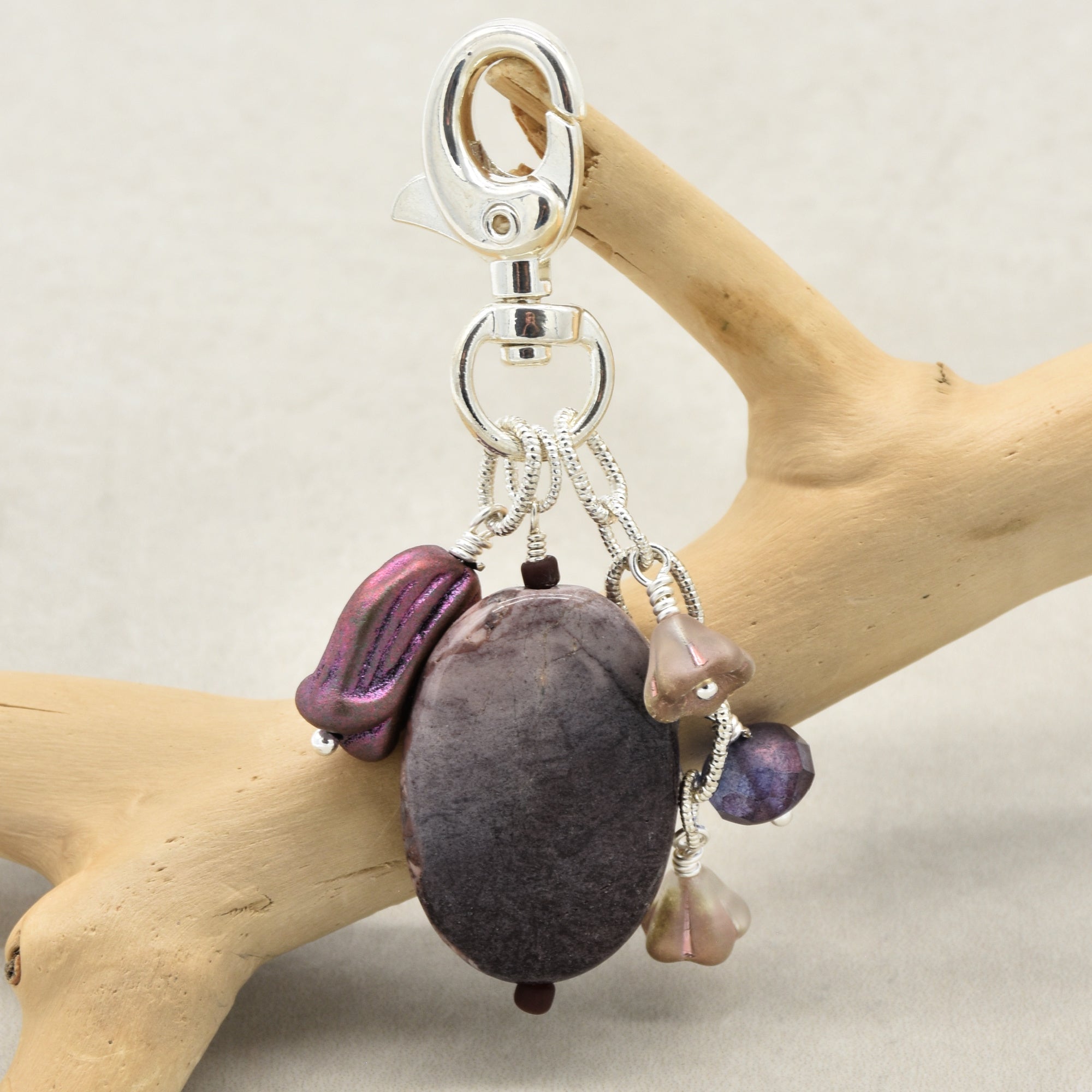 A handmade keychain featuring an oval porcelain jasper focal, a metallic purple Czech glass tulip charm, a small taupe flower charms, and a purple bead, all attached to a silver lobster clasp and displayed on a light wood branch against a neutral beige background - made with love in Colorado by Ruby Acorn.