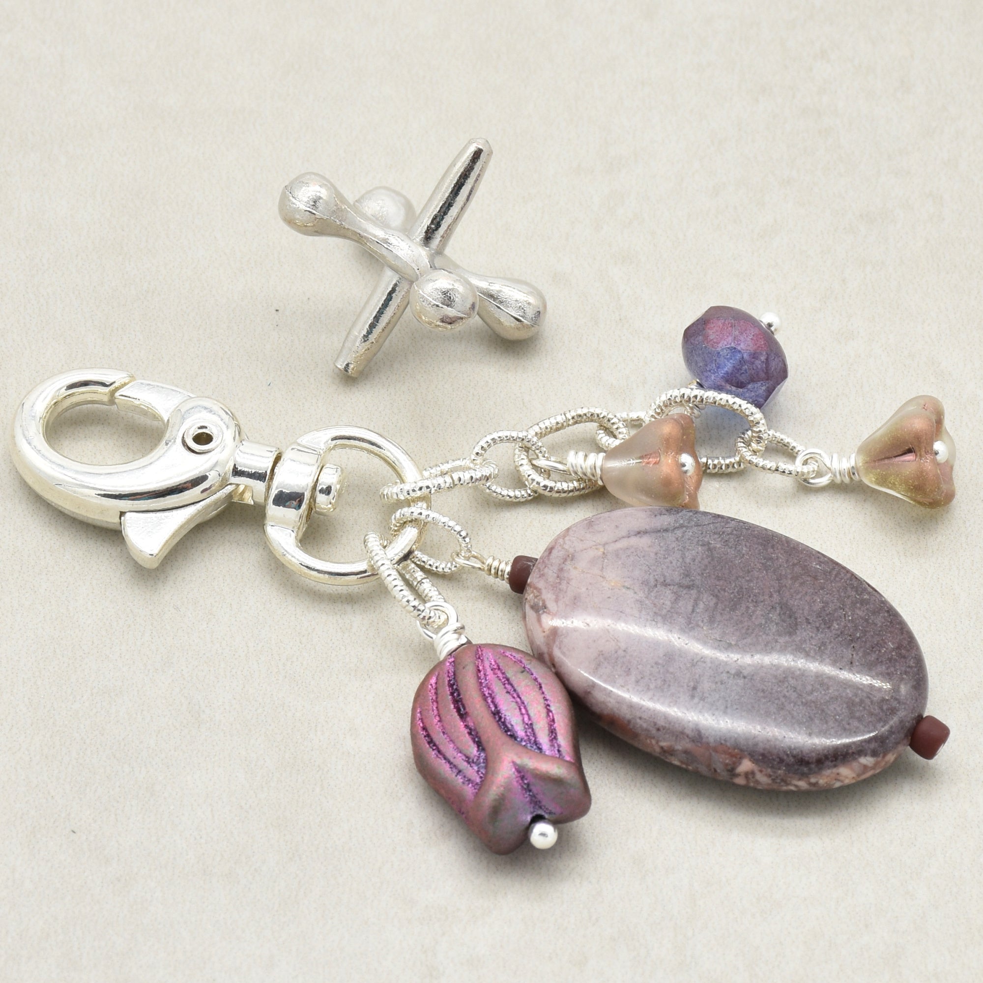 A silver-toned keychain or bag charm with a lobster clasp, featuring a large oval purple stone, two smaller iridescent purple beads, and three small metallic flower-shaped beads on a neutral beige background - made with love in Colorado by Ruby Acorn.