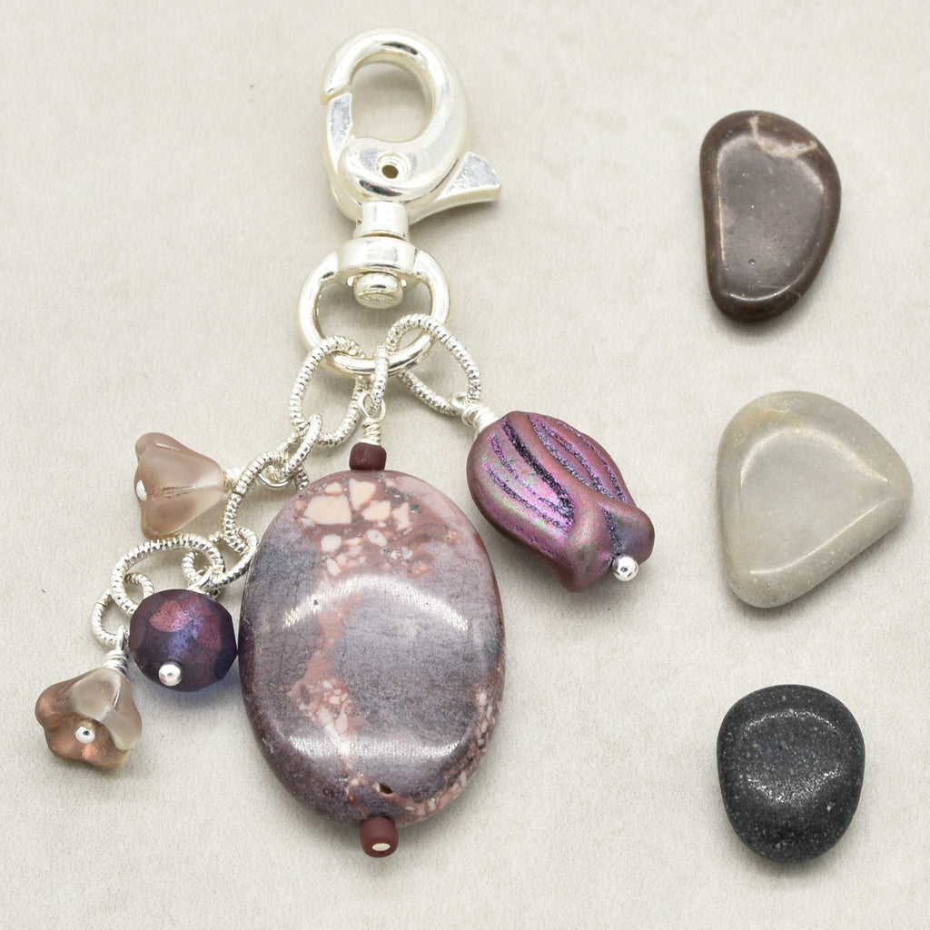 A silver keychain charm featuring an oval porcelain jasper focal, a purple iridescent bead, and several small glass beads, displayed next to three smooth decorative stones on a light beige background - made with love in Colorado by Ruby Acorn.