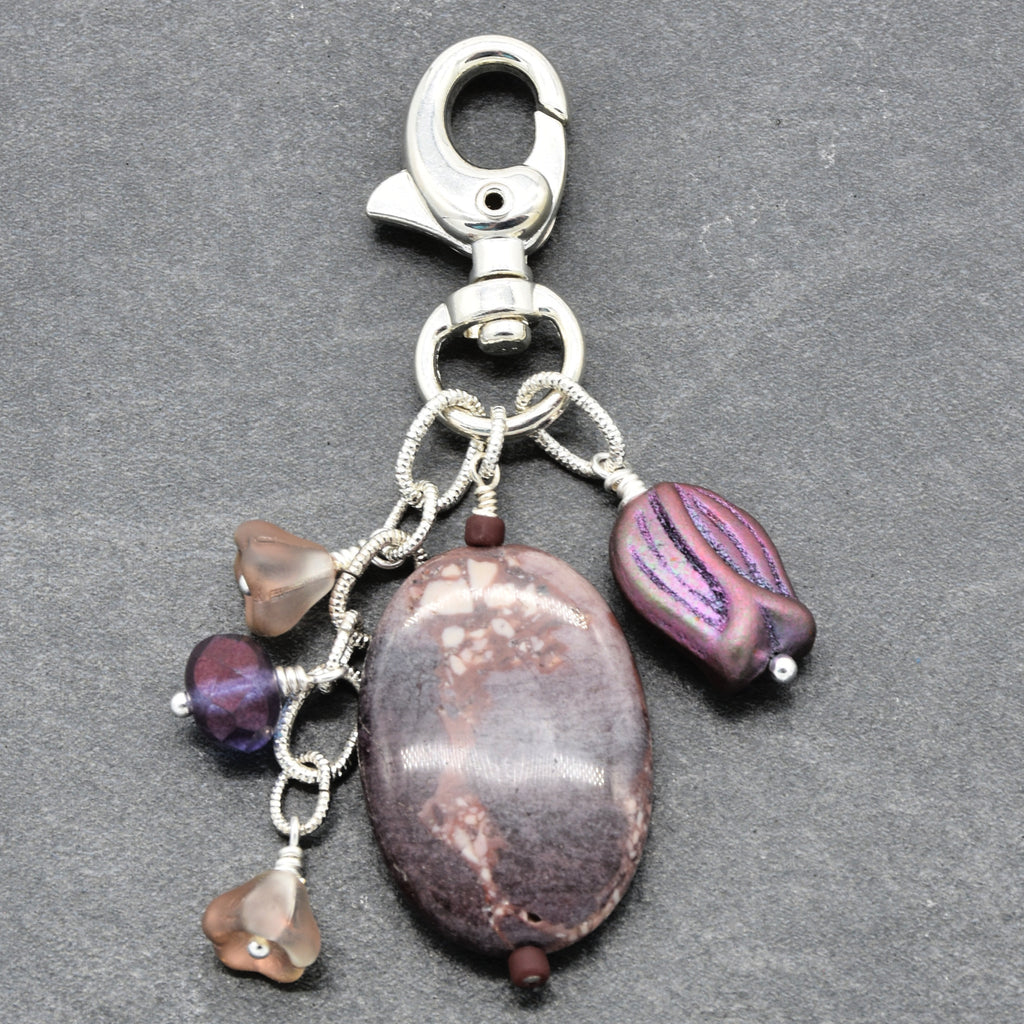 A silver-toned metal keychain clasp with multiple dangling beads and stones, including a large oval brown stone, a purple striped bead, and smaller translucent beads, shown on a dark gray textured background - made with love in Colorado by Ruby Acorn.