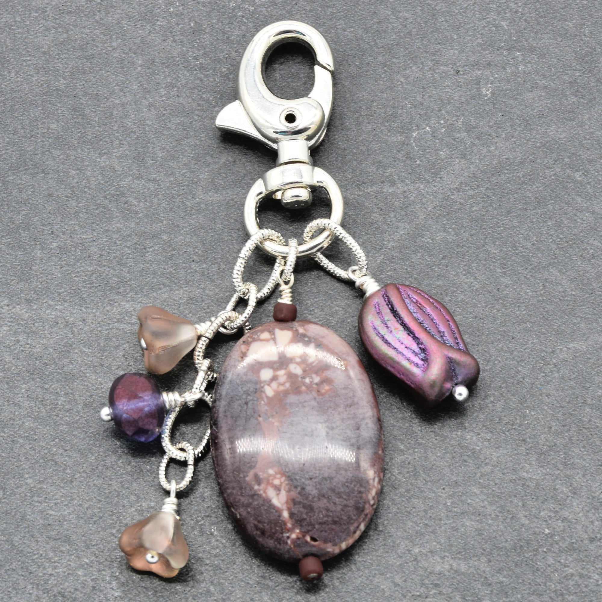 A silver-toned metal keychain clasp with multiple dangling beads and stones, including a large oval brown stone, a purple striped bead, and smaller translucent beads, shown on a dark gray textured background - made with love in Colorado by Ruby Acorn.
