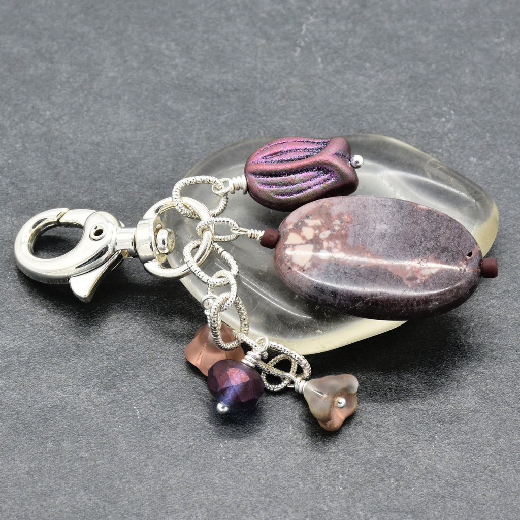 A handmade keychain or bag charm featuring a polished silver lobster clasp, an oval porcelain jasper focal, a smaller textured purple bead, and three dangling glass beads in purple and taupe, all arranged on a dark gray textured background - made with love in Colorado by Ruby Acorn.