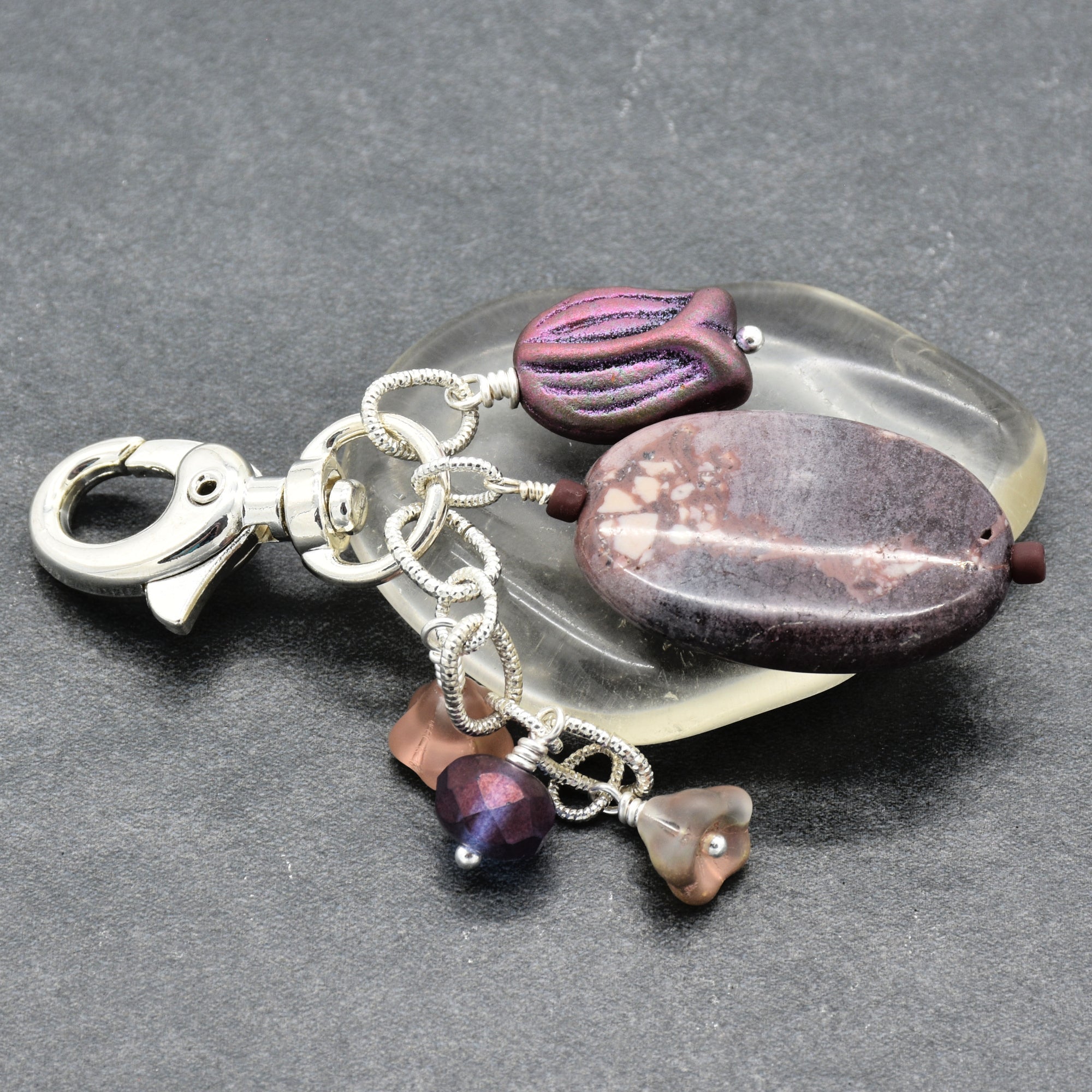 A handmade keychain or bag charm featuring a polished silver lobster clasp, an oval porcelain jasper focal, a smaller textured purple bead, and three dangling glass beads in purple and taupe, all arranged on a dark gray textured background - made with love in Colorado by Ruby Acorn.
