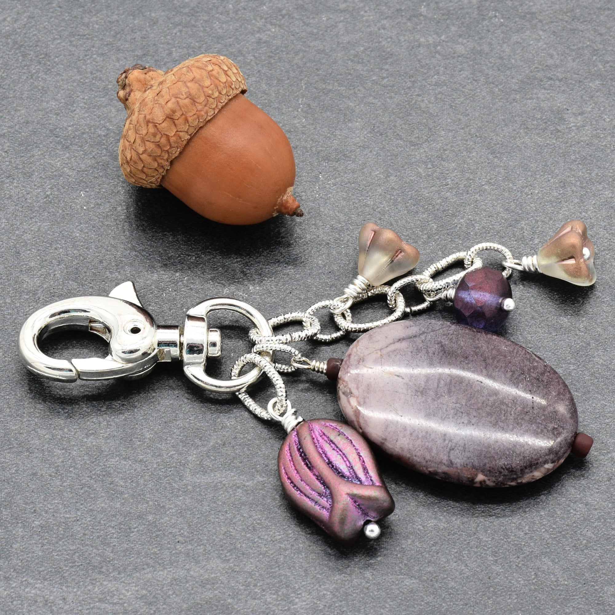 A handmade keychain featuring a silver lobster clasp, chain links, a porcelain jasper oval focal, a purple patterned bead, and three small glass beads, displayed on a dark gray textured background next to a decorative acorn - made with love in Colorado by Ruby Acorn.