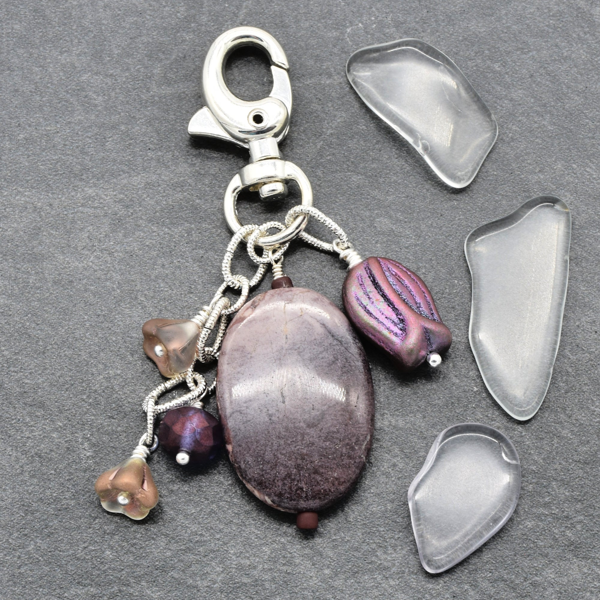 A handmade keychain charm featuring am oval porcelain jasper focal, a smaller purple bead with iridescent purple and pink highlights, three small glass bead charms, and a silver lobster clasp. It is displayed on a dark gray slate background alongside four clear, flat, irregular-shaped glass chips - made with love in Colorado by Ruby Acorn.