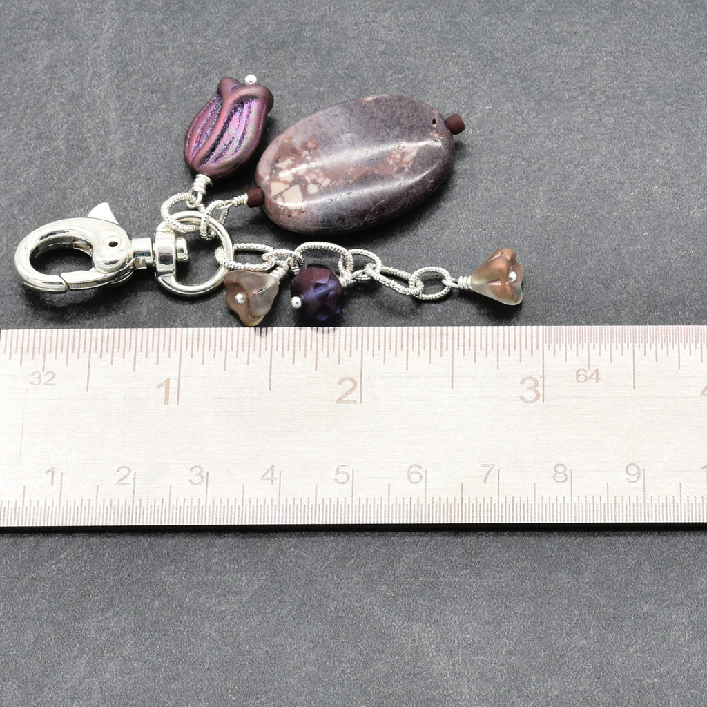 A handmade keychain or bag charm with purple and brown beads, a silver clasp, and a silver chain, shown next to a ruler on a dark gray background - made with love in Colorado by Ruby Acorn.