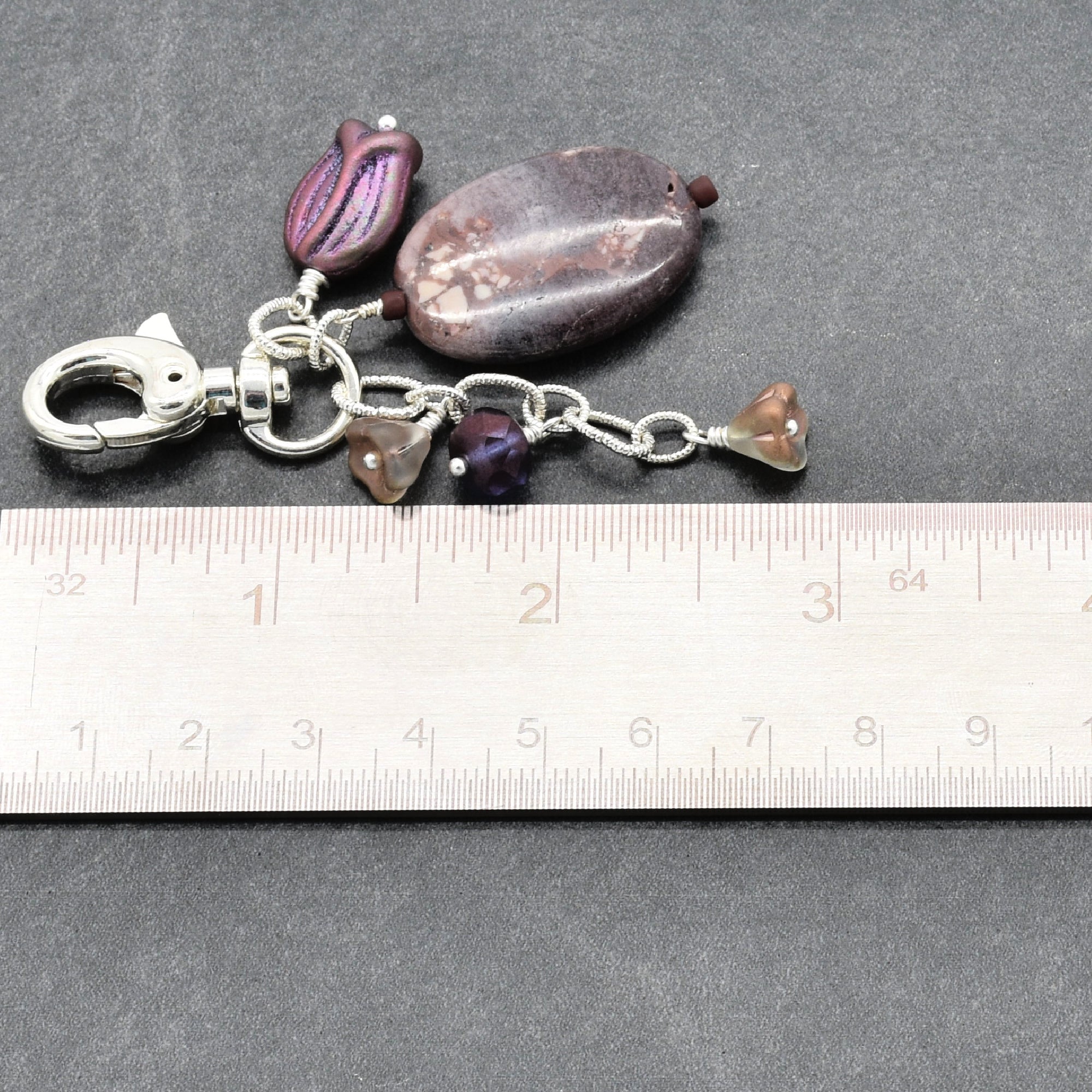 A handmade keychain or bag charm with purple and brown beads, a silver clasp, and a silver chain, shown next to a ruler on a dark gray background - made with love in Colorado by Ruby Acorn.
