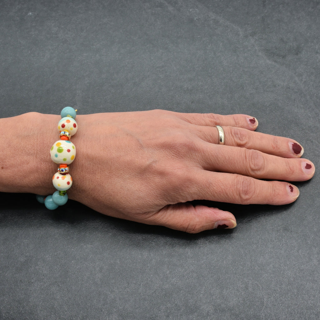 A close-up of a person's hand wearing a beaded bracelet. The bracelet features light blue beads, white beads with colorful polka dots, and small orange spacer beads.