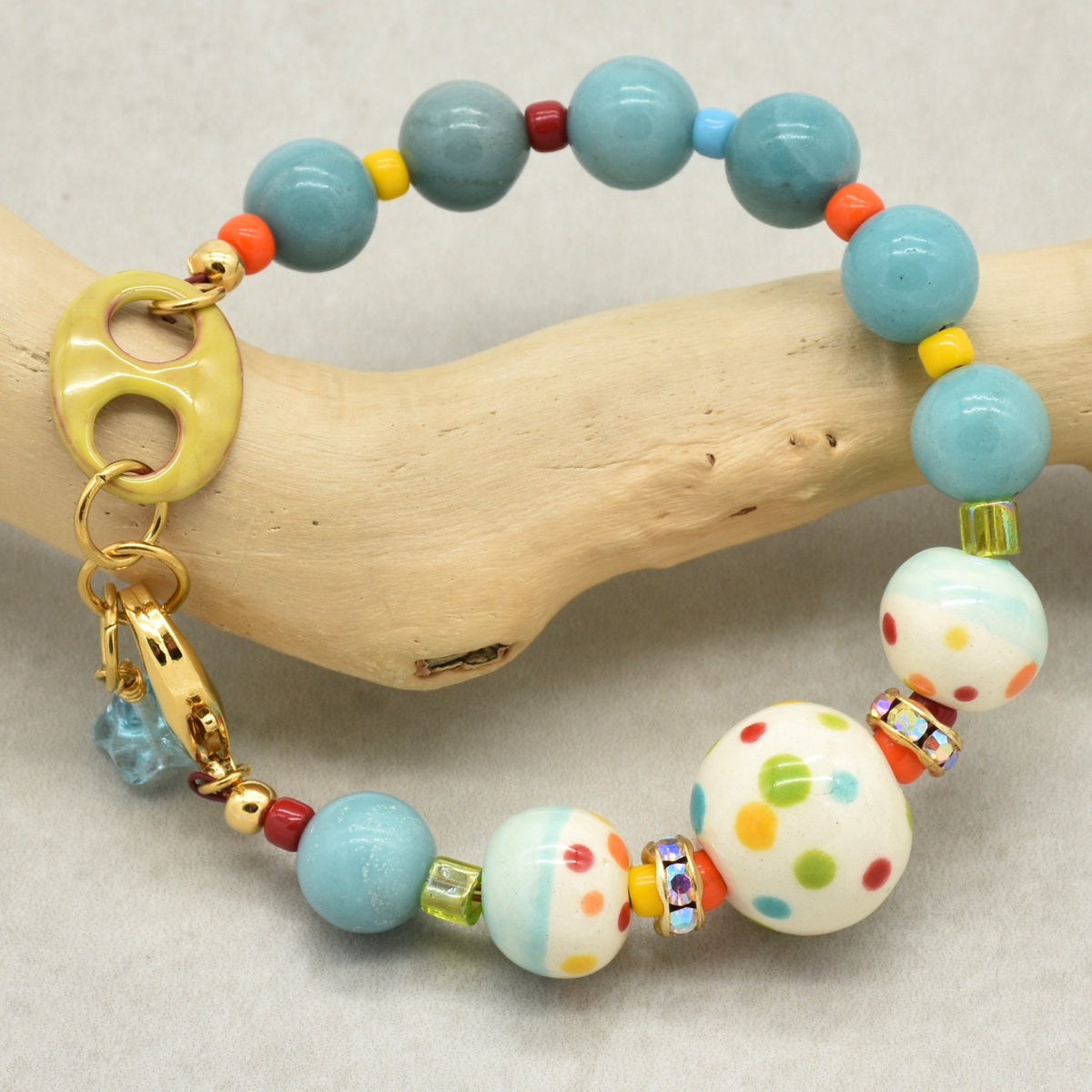 A colorful beaded bracelet displayed on a light wood branch. The bracelet features light blue marbled beads, three large white beads with multicolored polka dots, a yellow ring clasp, and small accent beads in red, orange, yellow, and green.