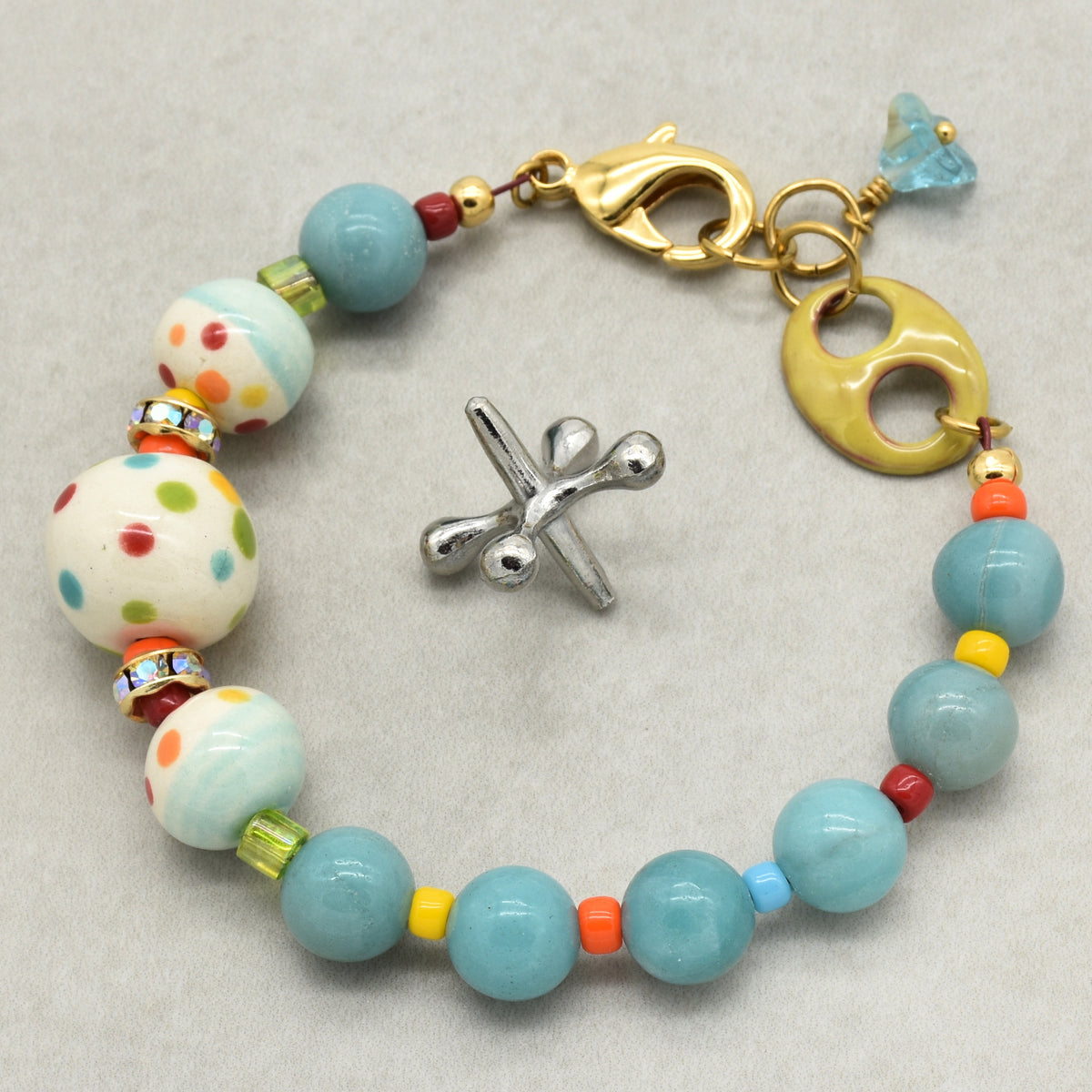 A handmade beaded bracelet with light blue, white, and multicolored beads, and gold-toned hardware, displayed on a neutral gray background.