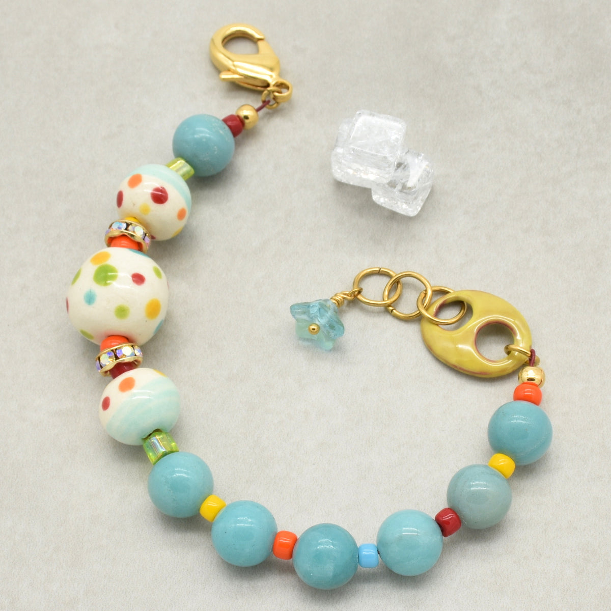 A handmade beaded bracelet with light blue, white, and multicolored beads, gold-toned hardware, and a small blue flower charm, displayed on a neutral gray background.