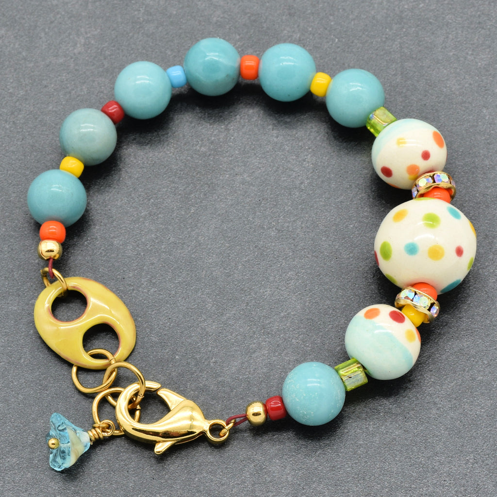 A colorful beaded bracelet with light blue, white polka-dot, and yellow beads, a gold-tone lobster clasp, and a small blue glass flower charm, displayed on a dark gray textured background.