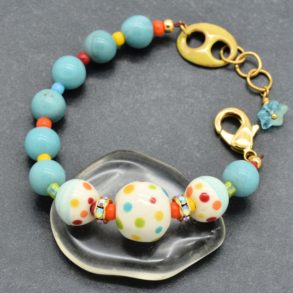 A handmade beaded bracelet with light blue amazonite beads, white polka-dot ceramic beads, and gold-toned metal hardware, displayed on a clear glass dish over a dark gray background.