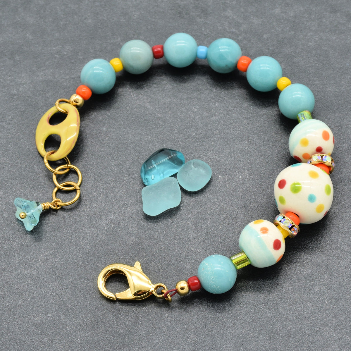 A handmade beaded bracelet with light blue amazonite beads, white beads with colorful polka dots, small orange and red beads, a yellow enameled link, and gold-tone clasp hardware, displayed on a dark gray background with three small blue glass chips.