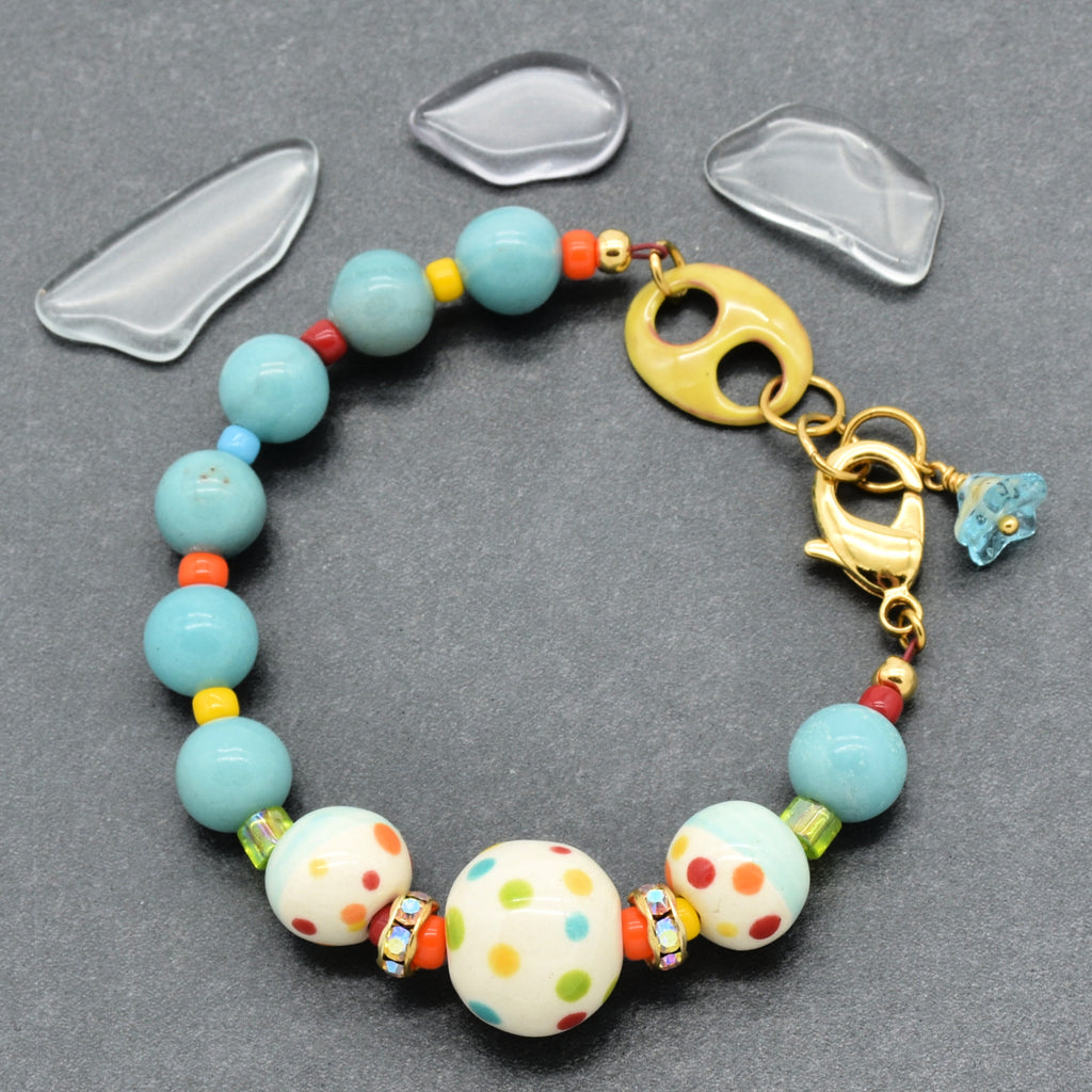 A handmade beaded bracelet with light blue, white polka-dot, and orange beads, a gold clasp, and a small blue flower charm, displayed on a dark gray background with three clear glass chips.
