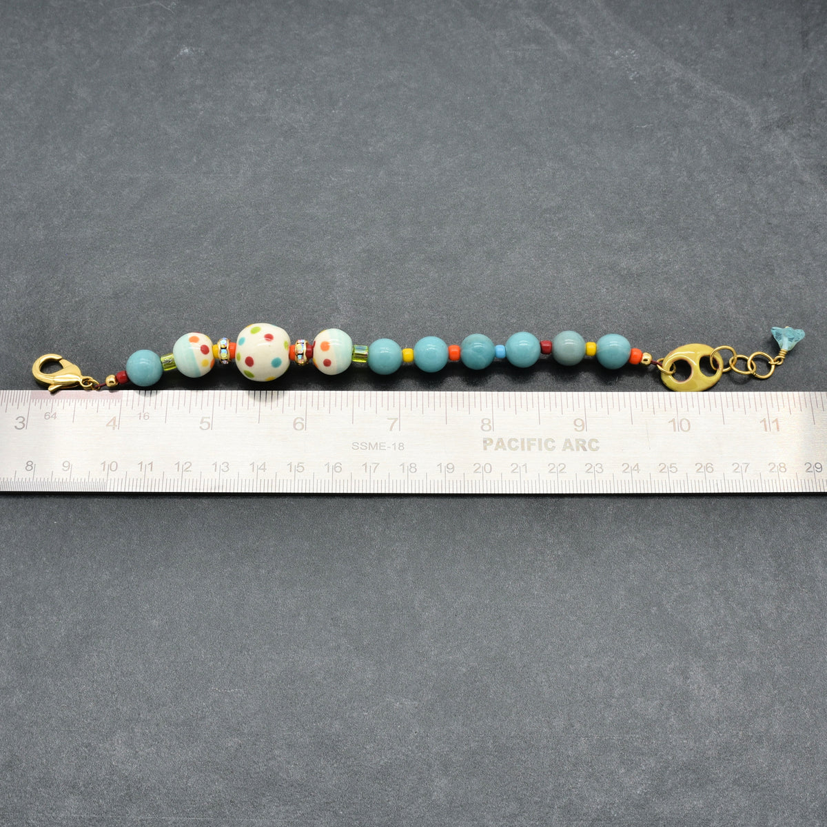 A handmade beaded bracelet with light blue, white, and red polka-dot beads, gold-toned hardware, and a small blue glass flower charm, shown next to a metal ruler for scale on a dark gray background.