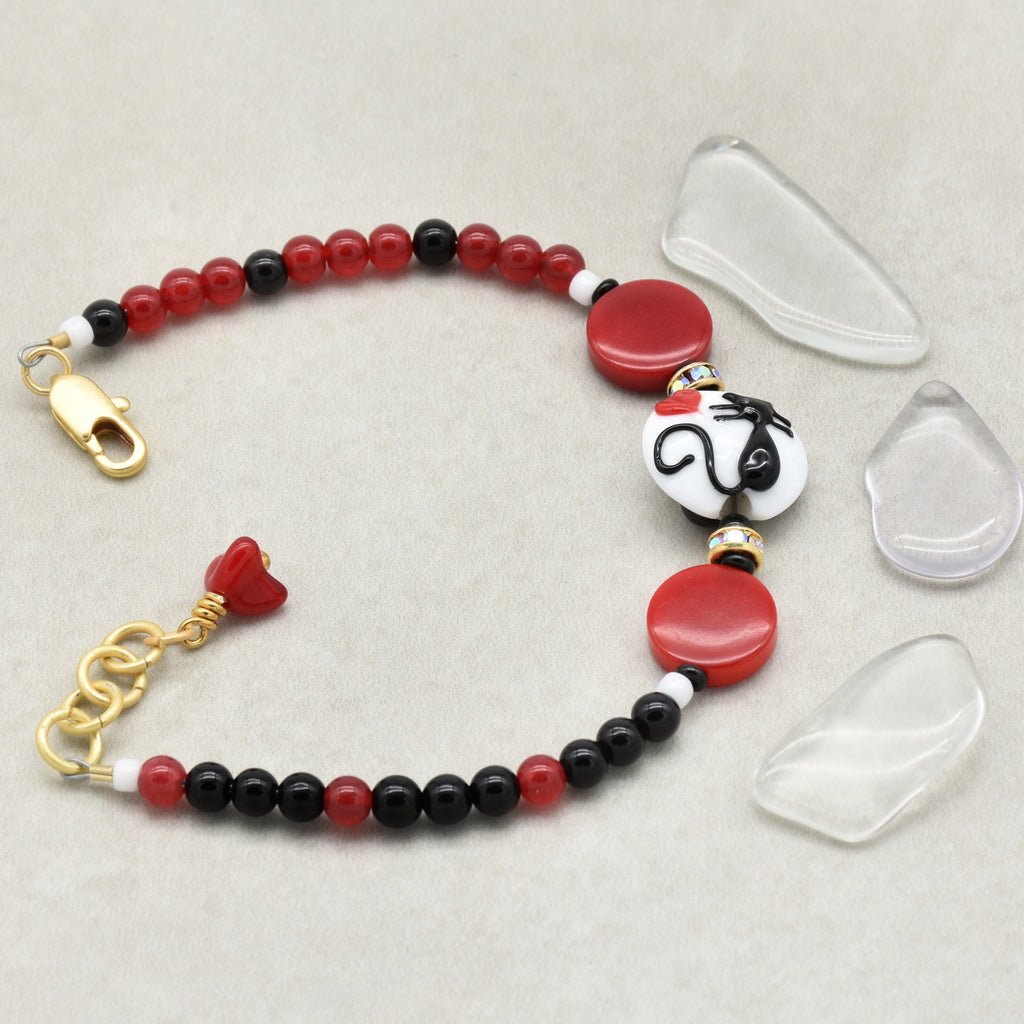 Beaded bracelet with red, black, and white beads, featuring a cat focal, on a light gray background.