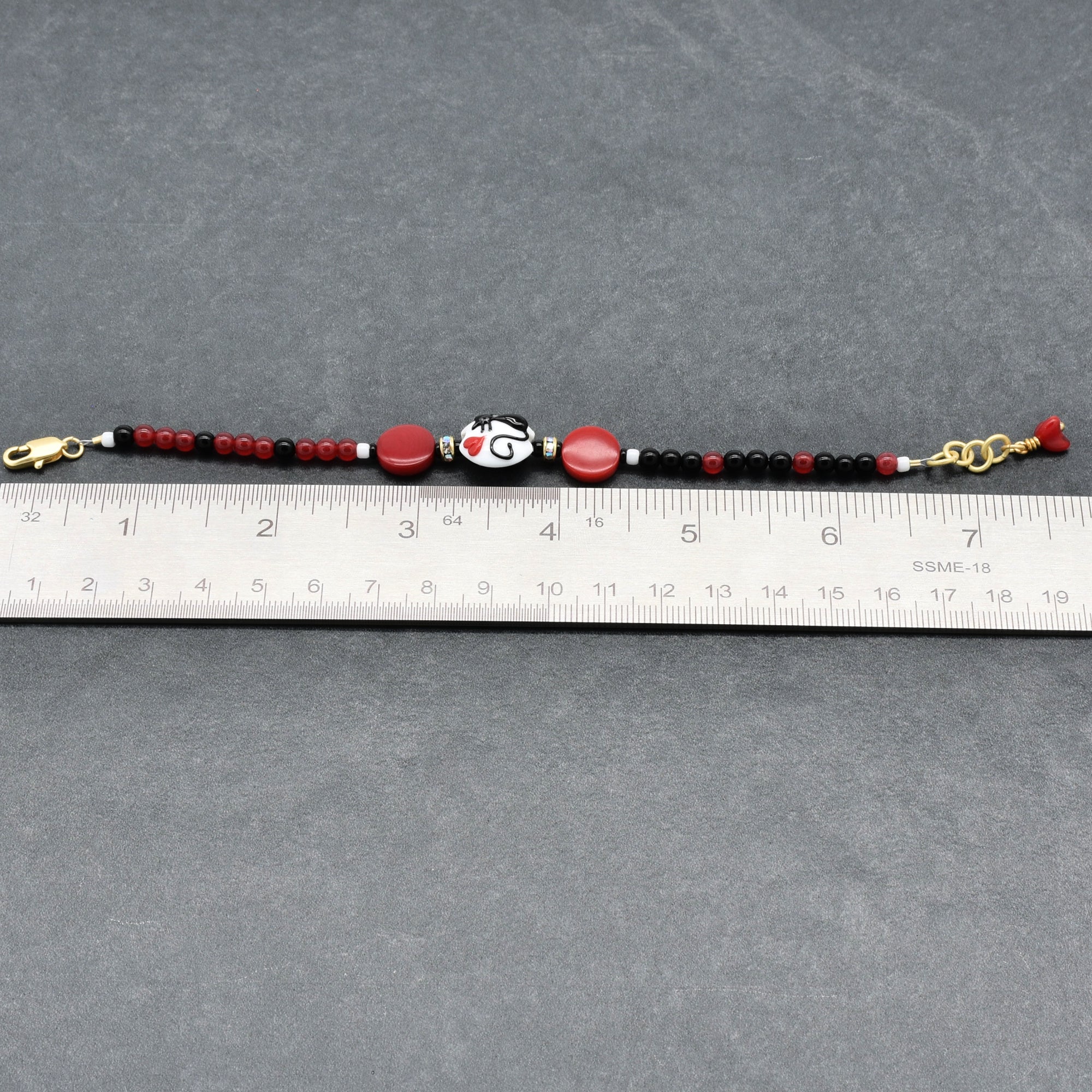 Beaded bracelet with red, black, and gold accents on a gray surface with a ruler for scale.