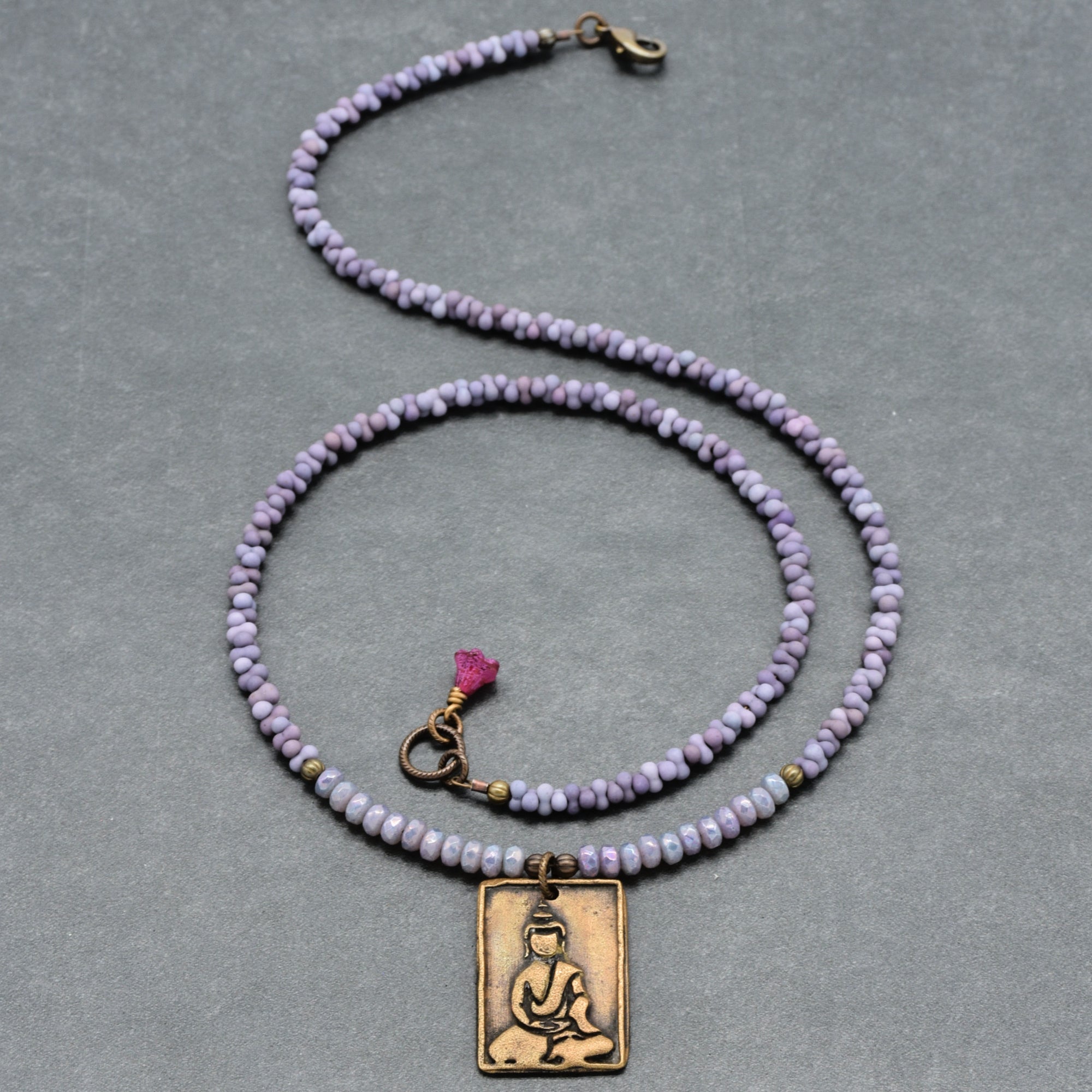 Purple beaded necklace with a brass Buddha pendant on a gray background