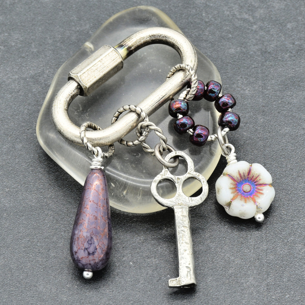 Keychain with various charms including a key, flower, and purple teardrop on a gray background