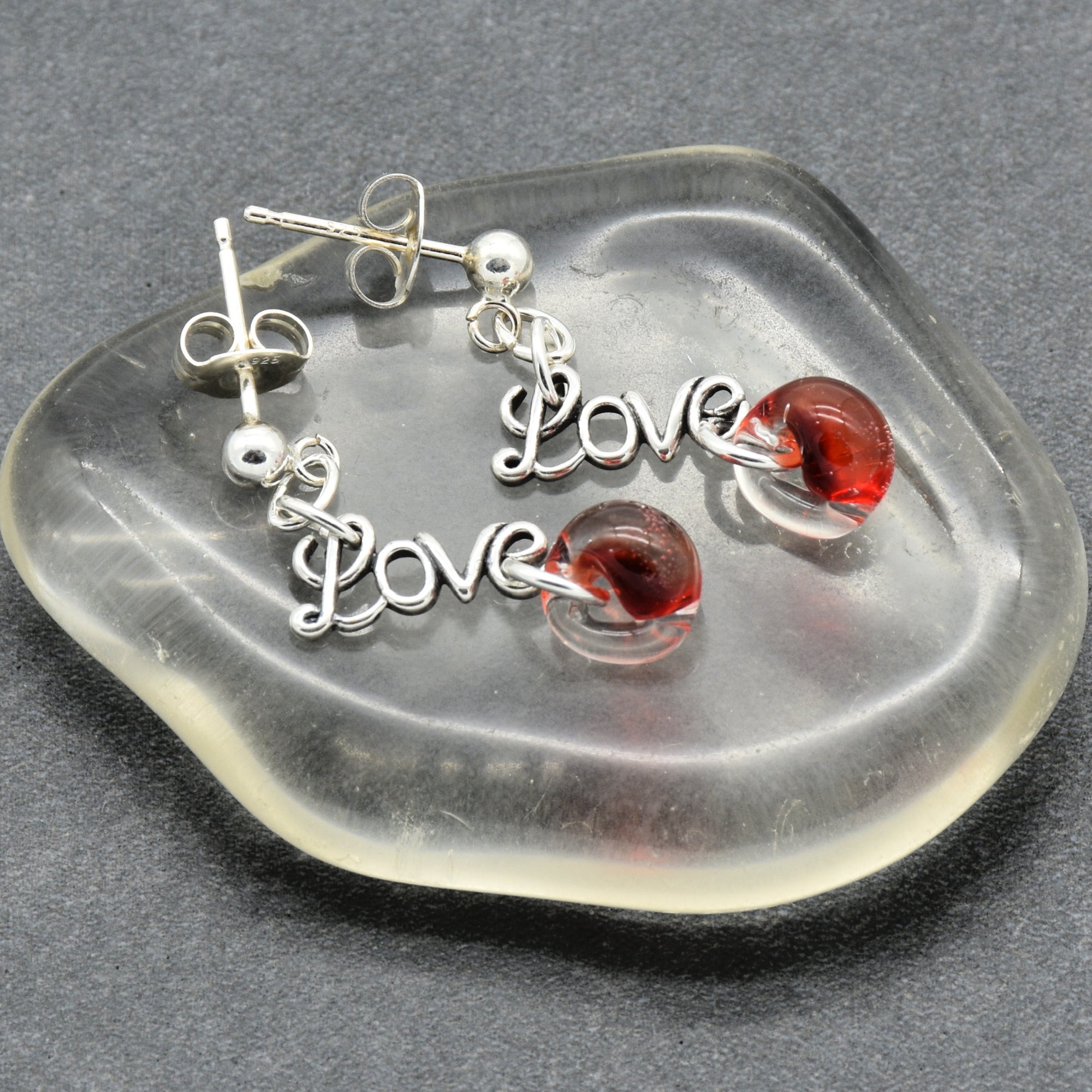Silver earrings with 'Love' charms and red glass teardrops on a clear stone surface