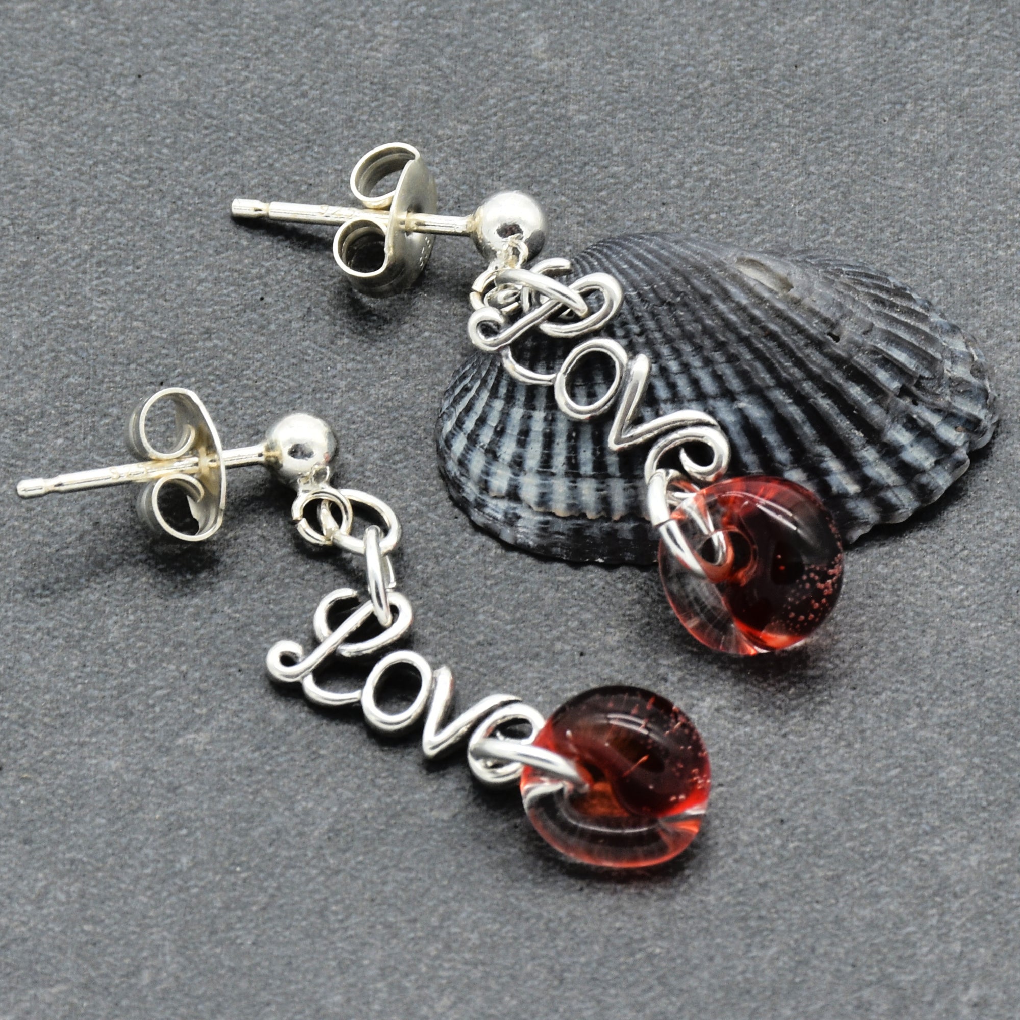 Silver earrings with 'LOVE' and red beads on a gray surface with a shell.