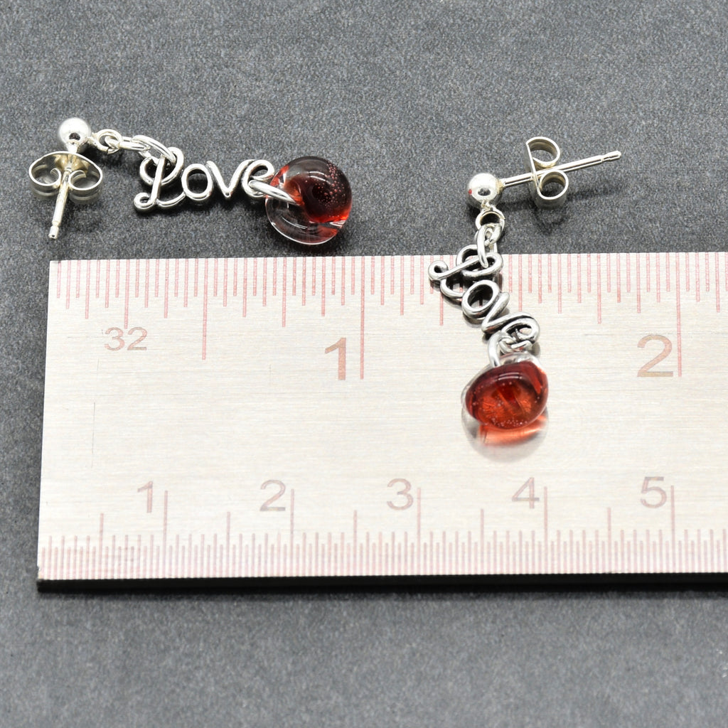 Pair of earrings with red charms and silver 'LOVE' charms, measured against a ruler.