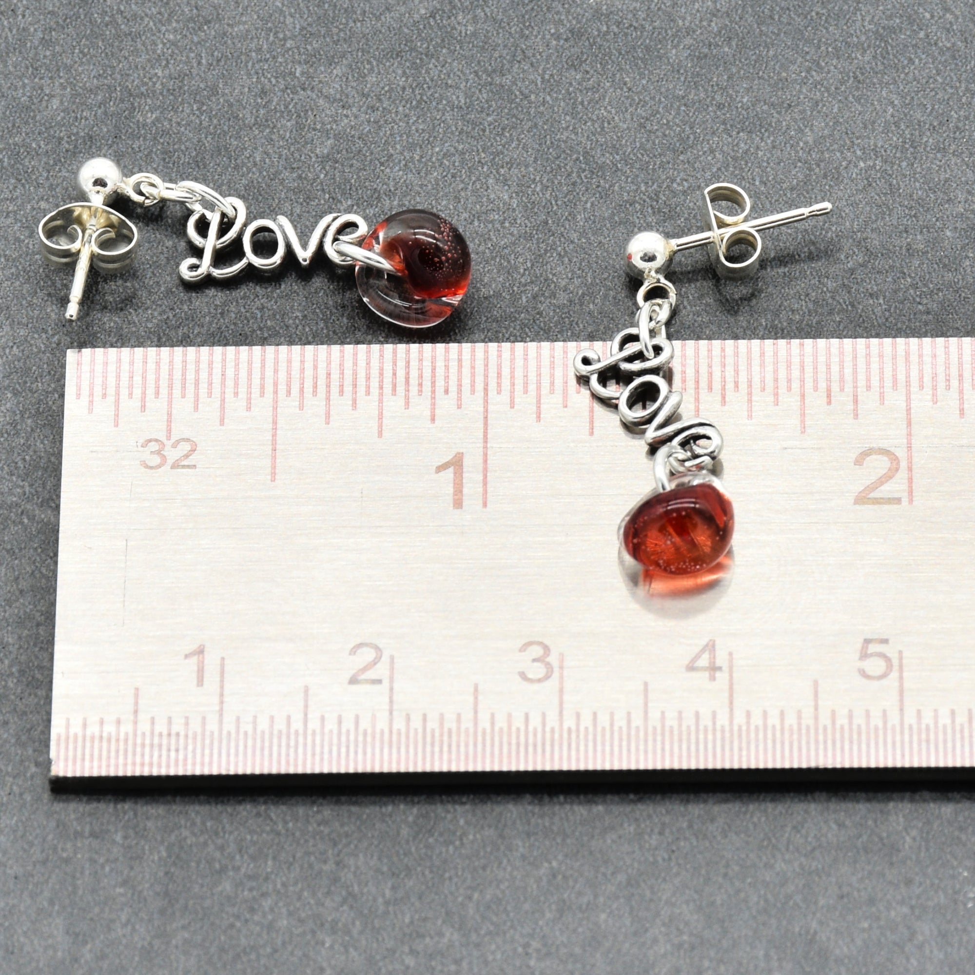 Pair of earrings with red charms and silver 'LOVE' charms, measured against a ruler.
