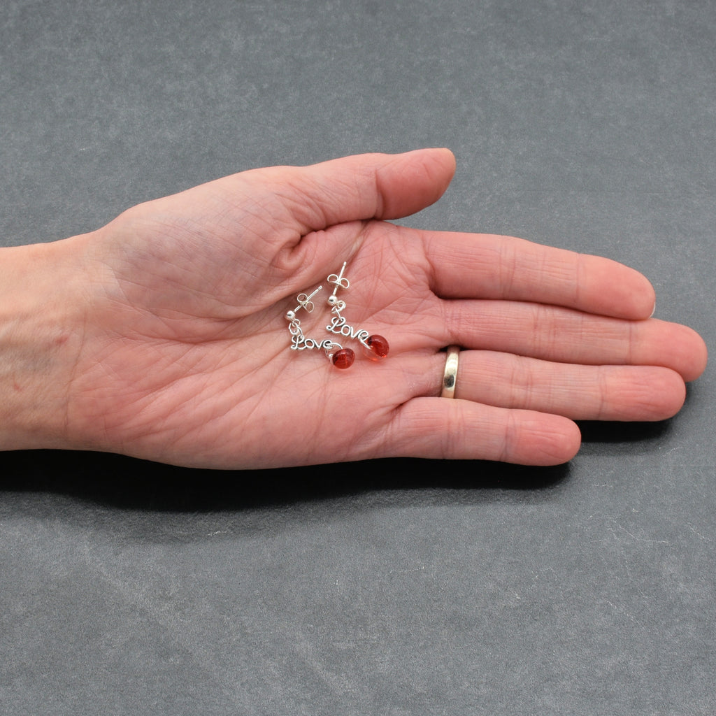 Hand holding silver earrings with red beads and 'love' charms on a gray background