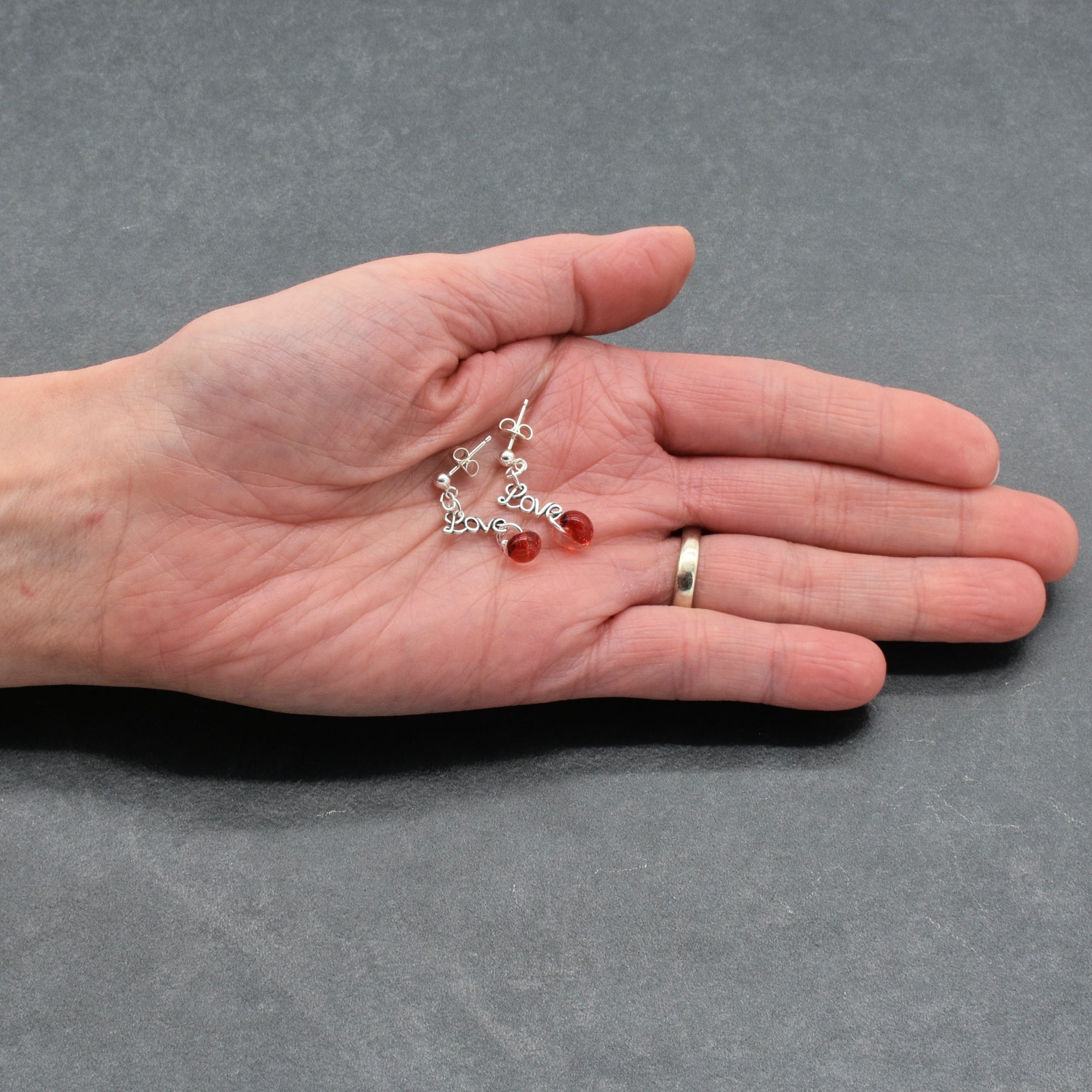Hand holding silver earrings with red beads and 'love' charms on a gray background