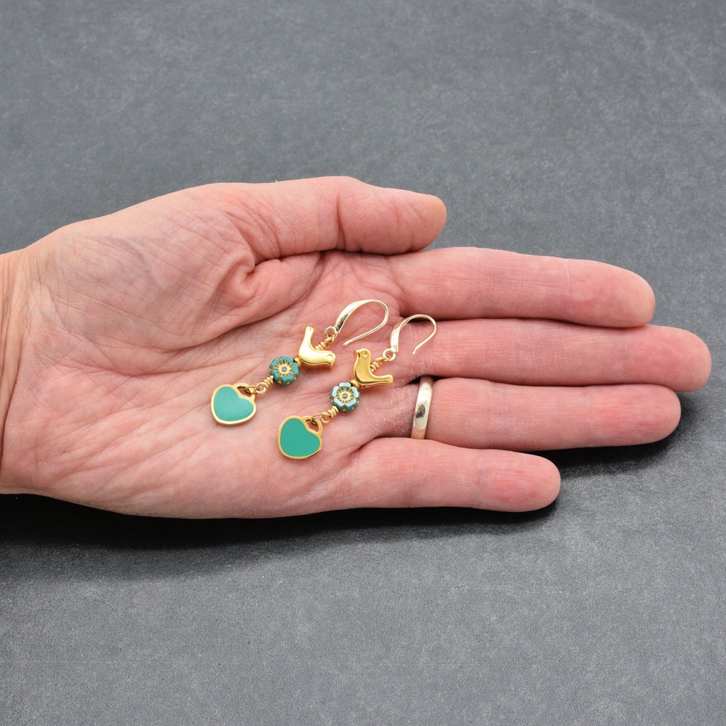 Artist's hand holding Lovebirds Turquoise Blue dangle earrings with gold birds and heart charms on a gray background - handmade with love by Ruby Acorn