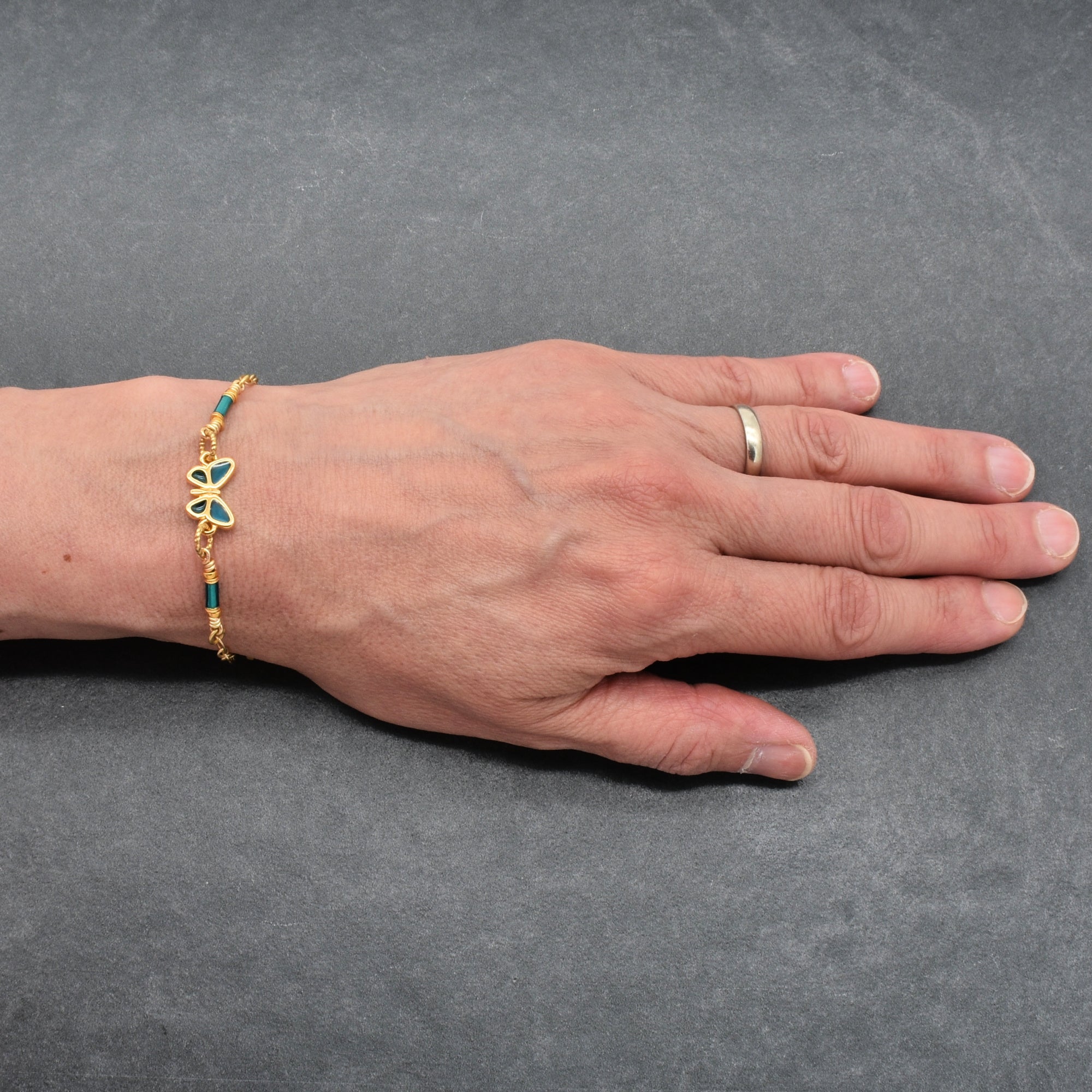 Hand wearing a gold bracelet with a blue butterfly charm on a gray background - wearable art by Ruby Acorn