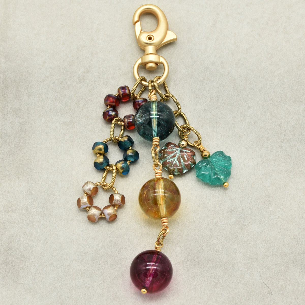 A gold-toned beaded keychain charm with multiple dangling beads and charms in green, red, brown, and turquoise, photographed on a light gray textured background.
