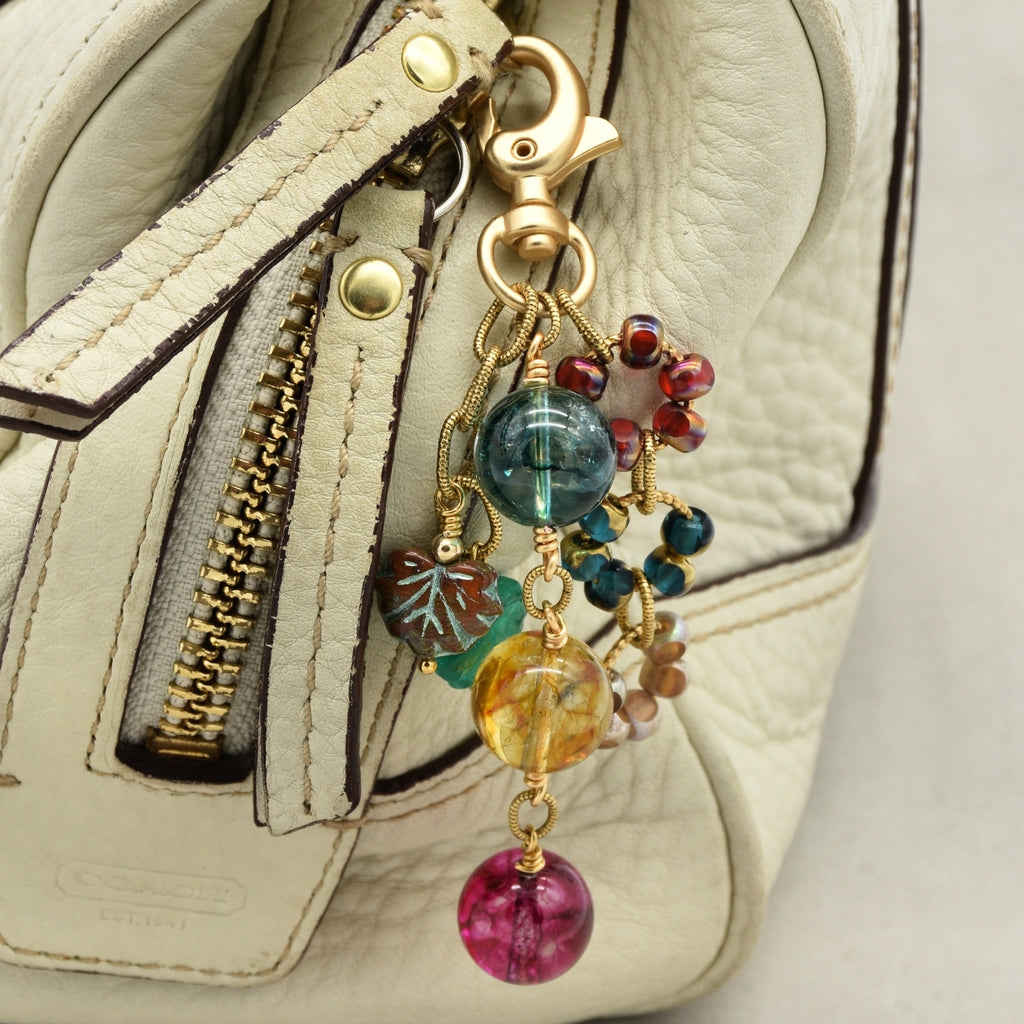 A close-up of a cream-colored leather handbag with a gold-tone clasp and a colorful beaded keychain charm featuring marbled glass beads in blue, yellow, pink, and teal.