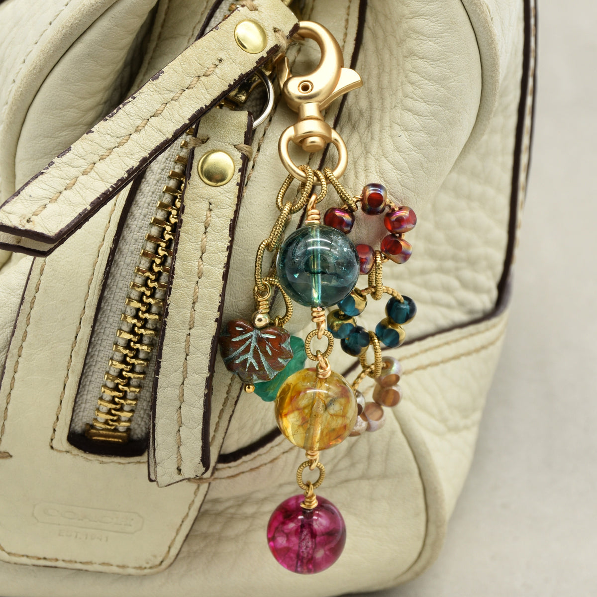 A close-up of a cream-colored leather handbag with a gold-tone clasp and a colorful beaded keychain charm featuring marbled glass beads in blue, yellow, pink, and teal.