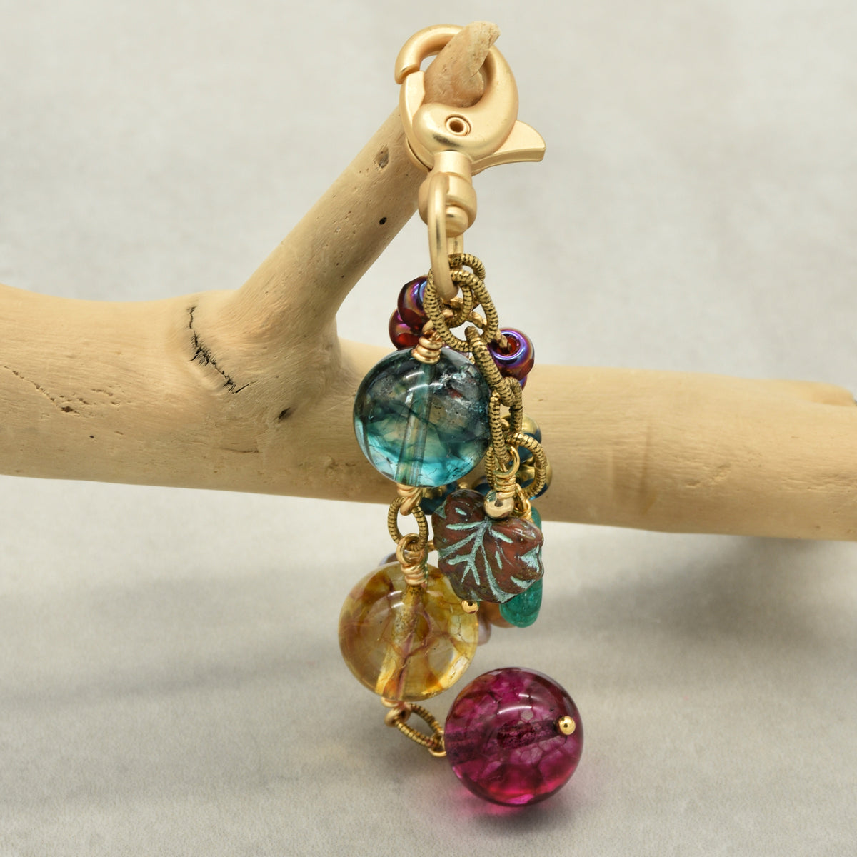 A handmade beaded keychain or bag charm with a gold-tone lobster clasp, hanging from a light wood branch. The charm features multiple colorful beads in teal, yellow, magenta, and purple, along with a small green leaf bead and gold wire details, set against a neutral gray background.