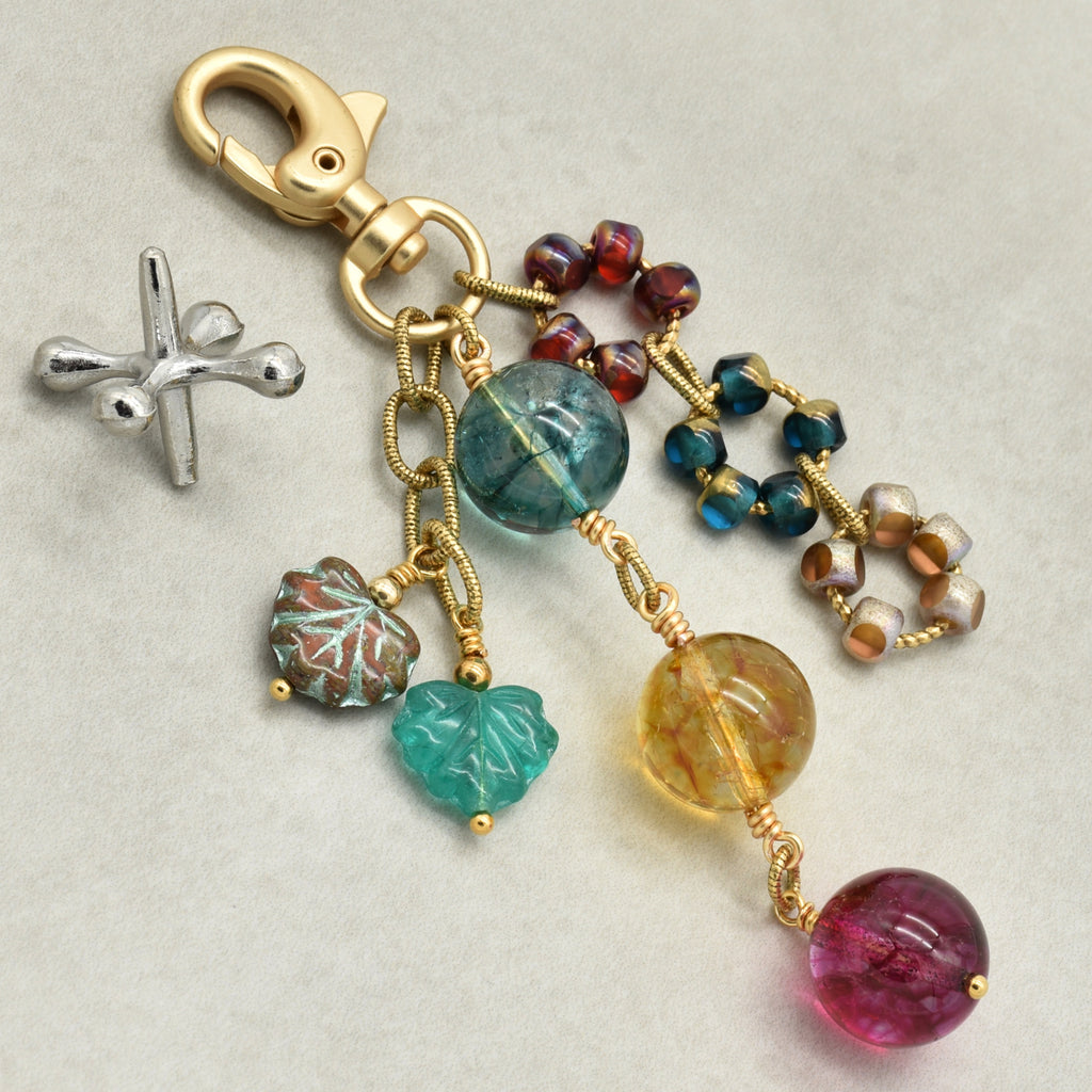 A gold-toned keychain or bag charm with a lobster clasp, featuring multiple dangling charms including a marbled green and white leaf charm, a turquoise leaf charm, a large round aqua bead, a yellow marbled bead, a cluster of blue beads, and a large round magenta bead, all displayed on a light gray background.