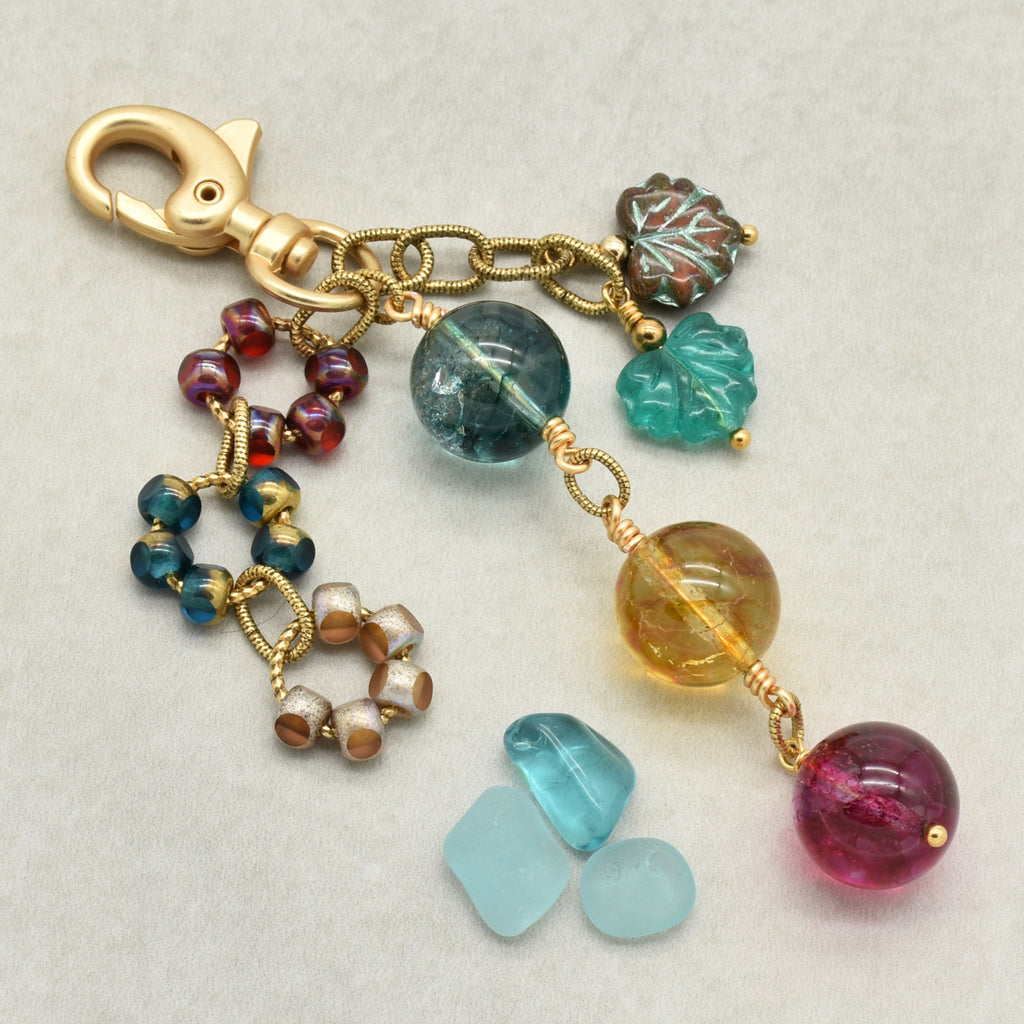 A handmade keychain charm featuring a gold-tone lobster clasp, chain, and multiple colorful beads and charms including a leaf charm, a blue-green round bead, a yellow translucent bead, and a pink round bead, displayed on a light gray textured background.