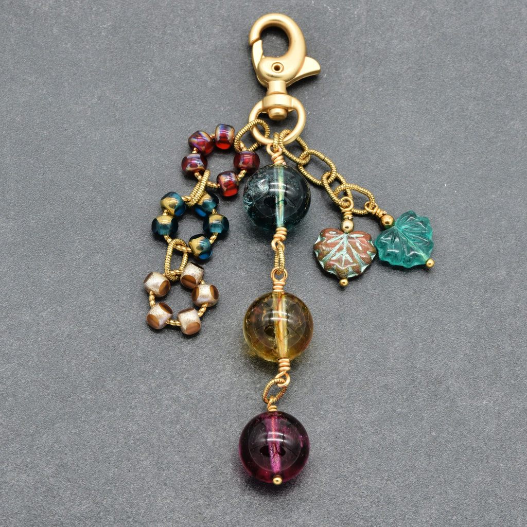 A gold-toned lobster clasp keychain charm featuring multiple dangling beaded and carved charms in green, purple, red, and gold, displayed on a dark gray textured background.