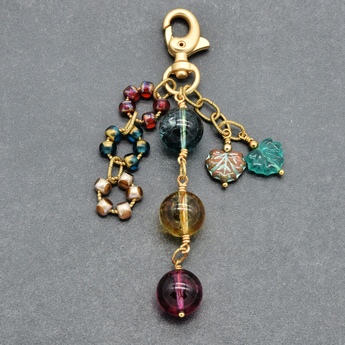 A gold-toned lobster clasp keychain charm featuring multiple dangling beaded and carved charms in green, purple, red, and gold, displayed on a dark gray textured background.