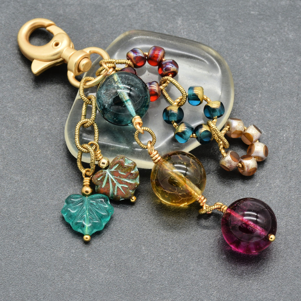 A gold-tone metal keychain or bag charm with a lobster clasp, featuring multiple colorful glass beads, wreathe-shaped beads, a green leaf charm, and a brown leaf charm, displayed on a clear stone stand over a dark gray textured background.